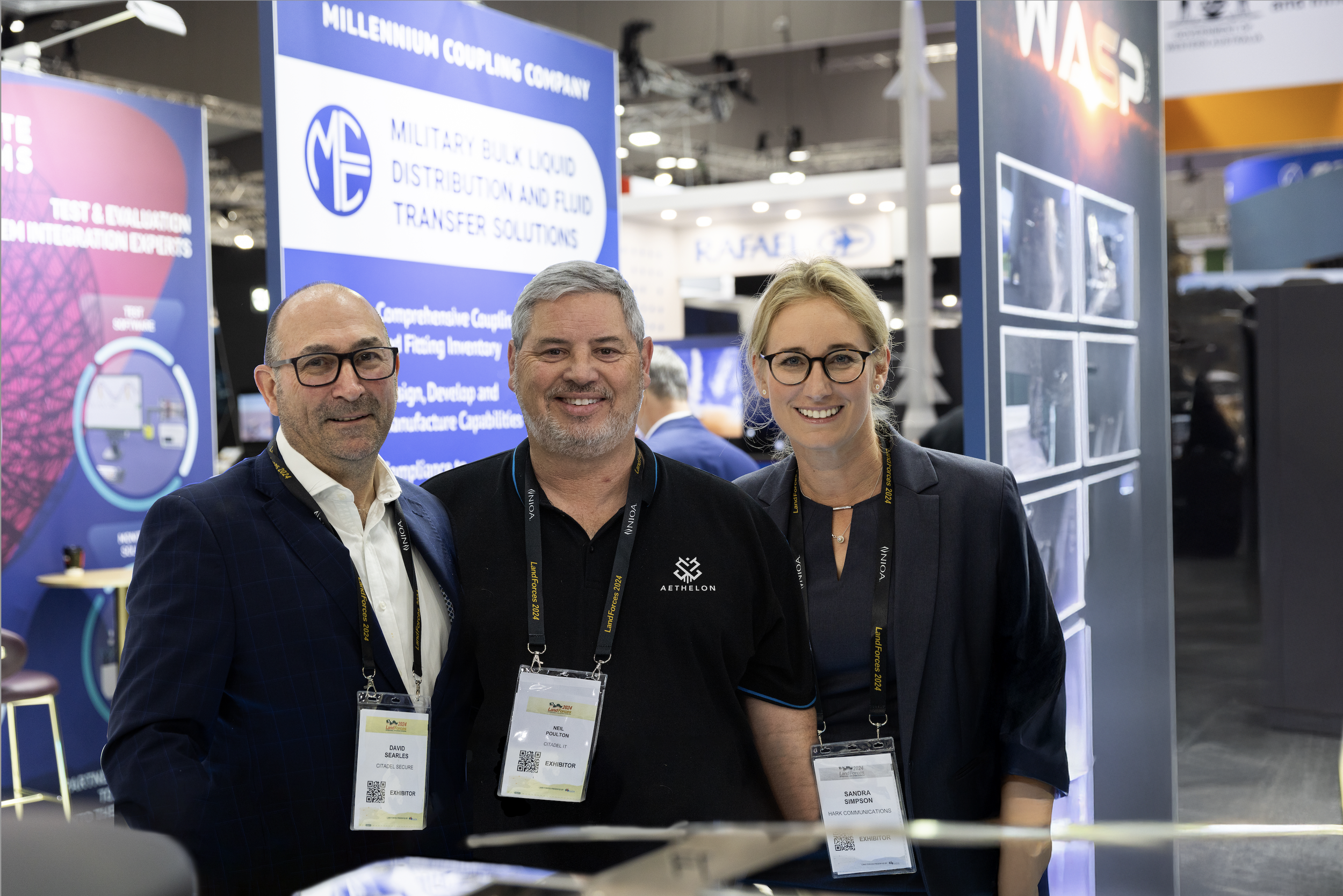 Three professionals, two men and one woman, standing in front of exhibition booths at a conference. All are wearing badge lanyards and smiling at the camera.