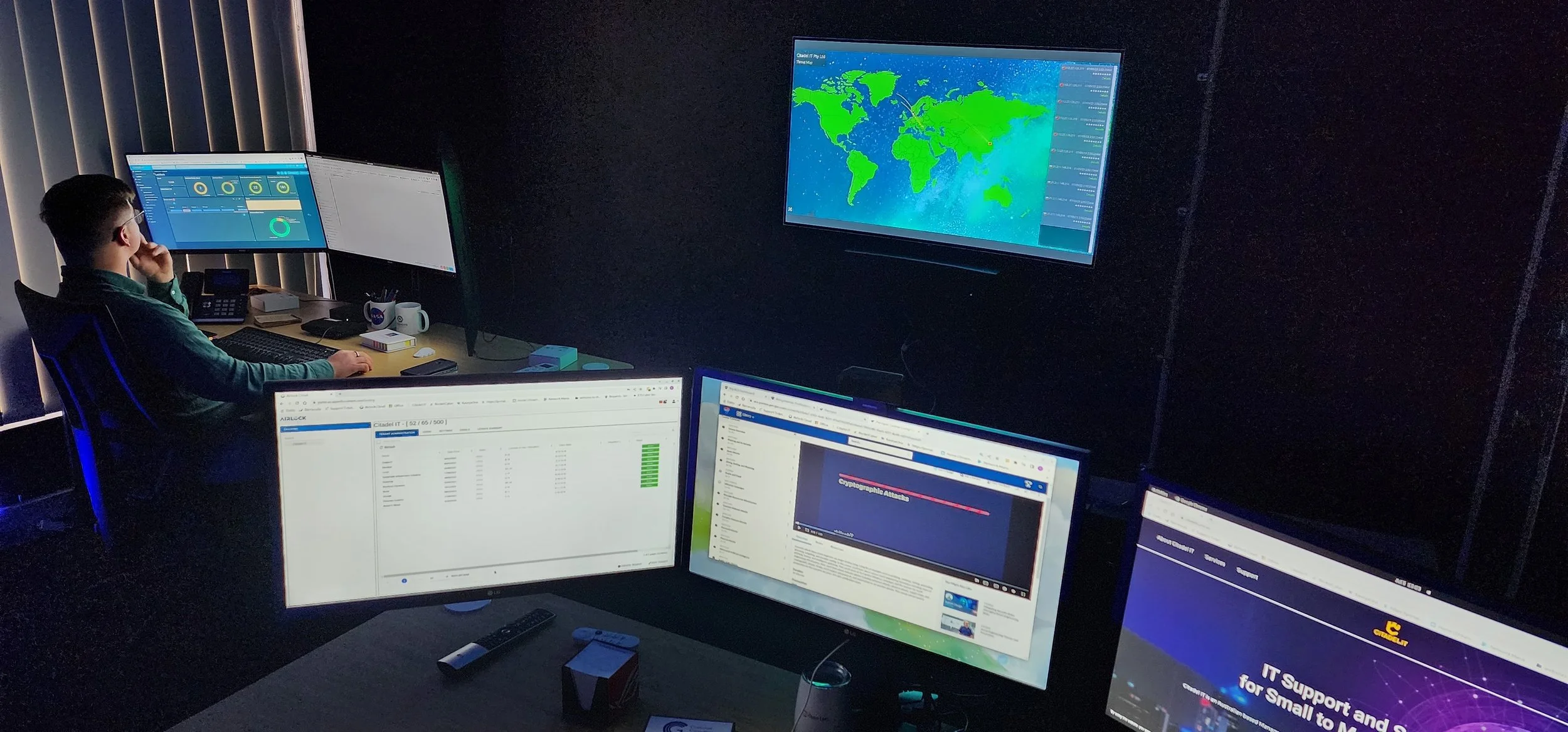 A person working in a dark office with multiple computer screens displaying world maps, data charts, and web pages.