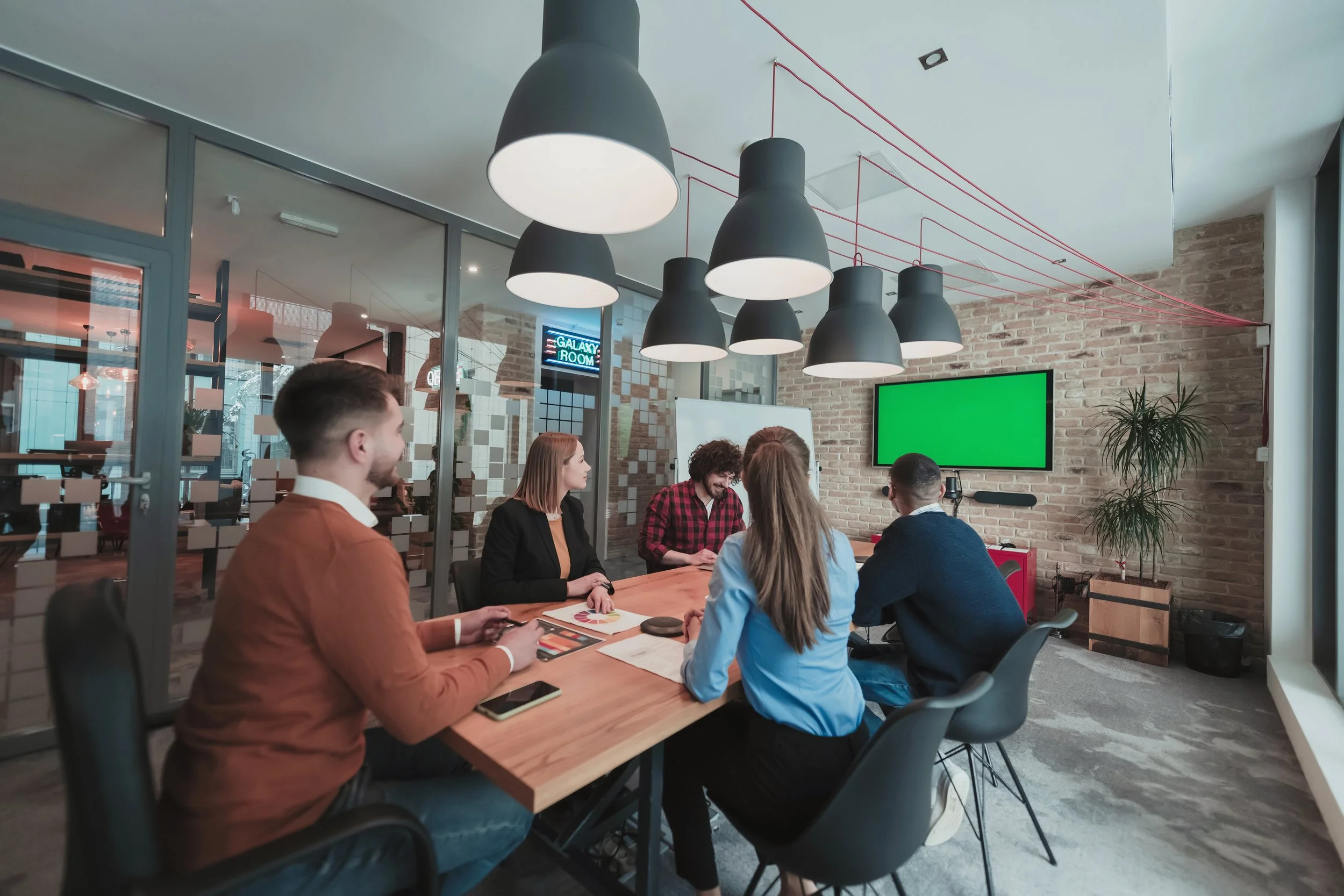 Group of five people in a meeting room, sitting around a wooden table, some with laptops and tablets, facing a green screen on the wall, in an office with exposed brick walls and modern black pendant lights.