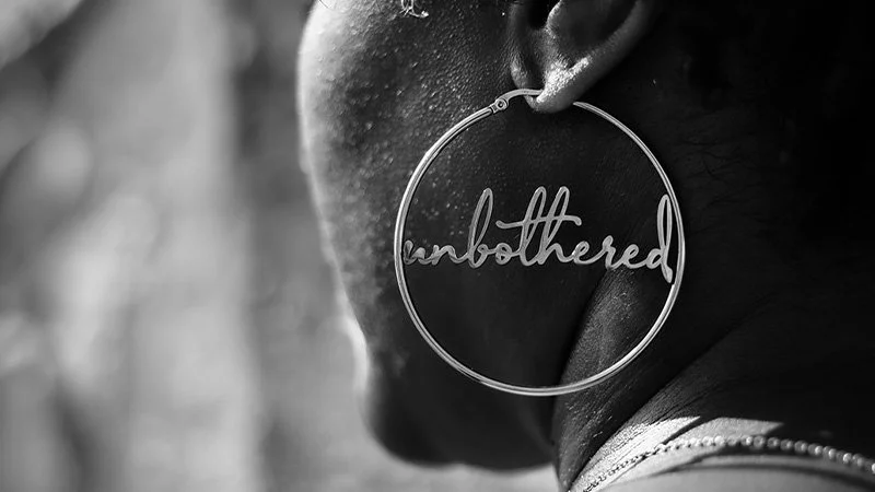 Close-up of a woman's earring with the word 'unbothered' in cursive inside a circular wire hoop