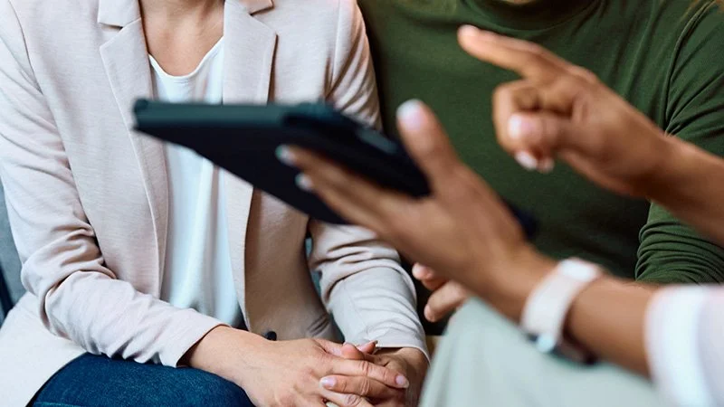 Two people engaged in a discussion, one showing a tablet and the other gesturing towards it, with a focus on their hands and partly visible torsos.