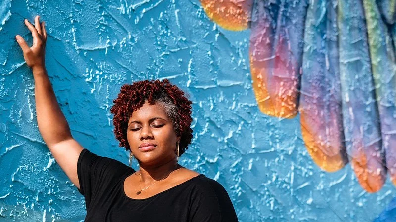 Woman with short curly hair standing against a textured bright blue wall with colorful decoration in background, eyes closed, one arm raised, appearing to be relaxing or meditating.