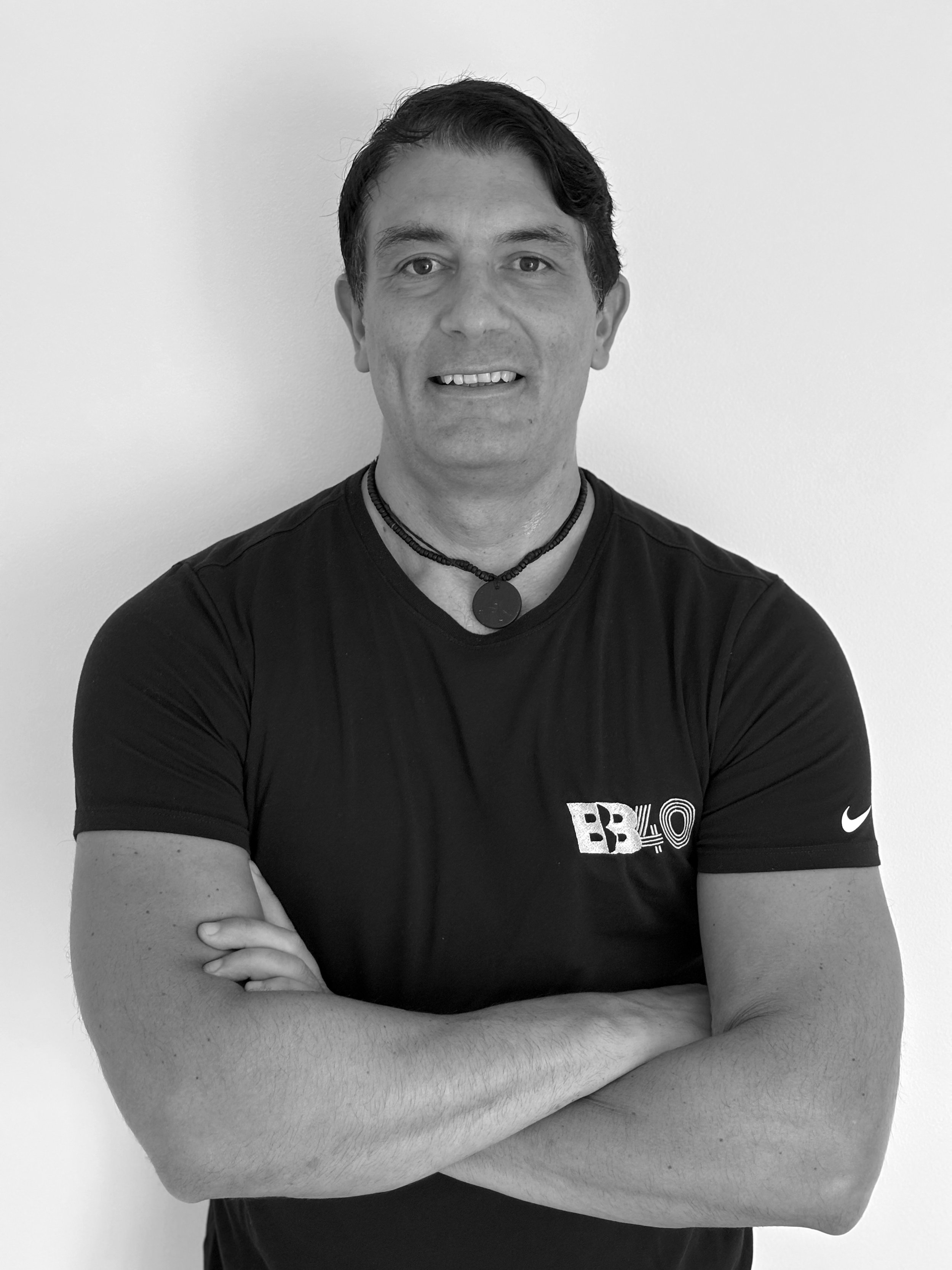 Black and white photo of a man with crossed arms, wearing a black T-shirt with logos, and a beaded necklace, standing against a plain wall.