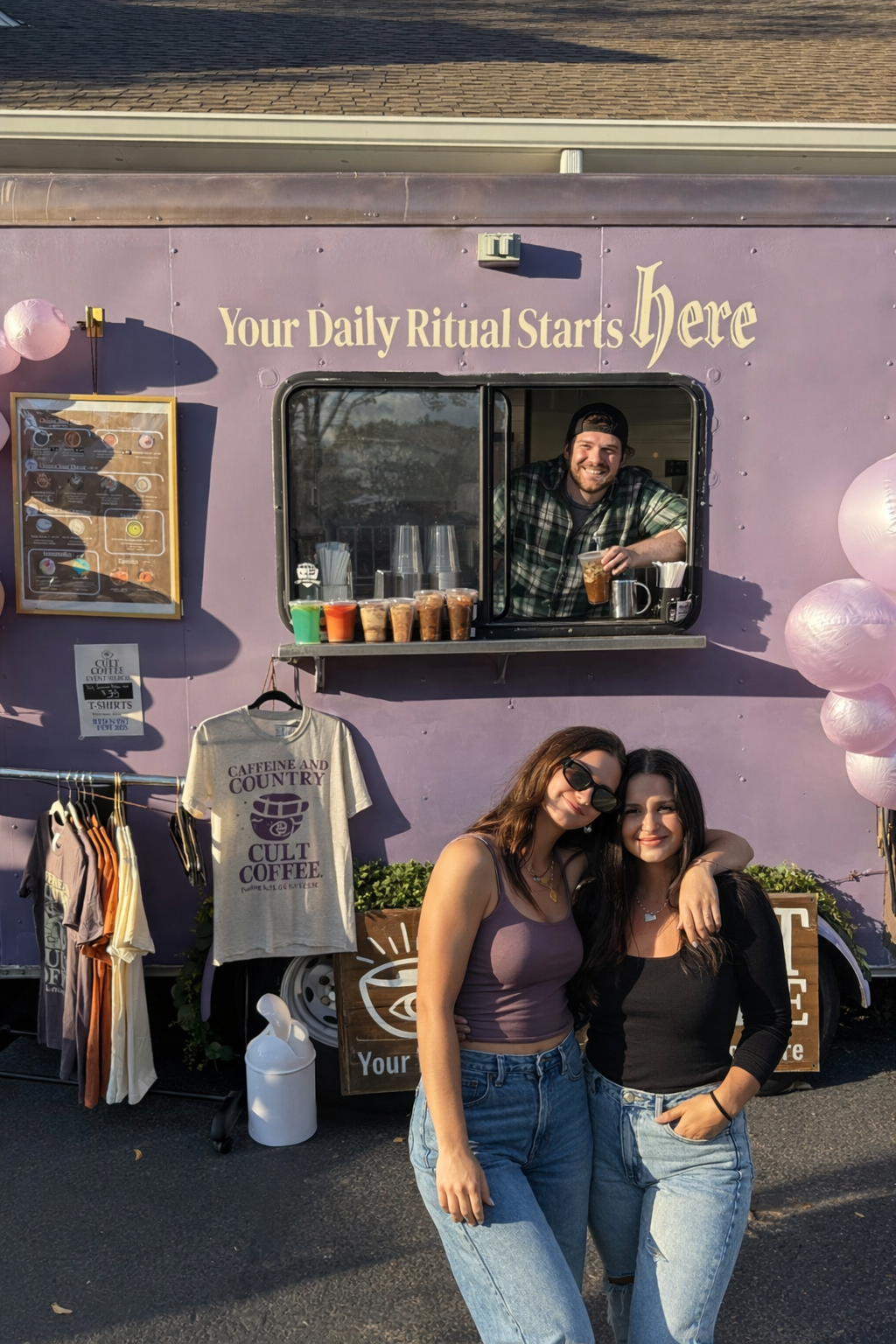 A lavender-colored food truck with the sign 'Your Daily Ritual Starts Here' and a window where a smiling man is serving drinks. Two women are standing in front of the truck posing for the photo, one with sunglasses and both smiling. There are t-shirts hanging outside, including one that says 'Caffeine and Country, Cult Coffee,' and balloons on the right side of the truck.