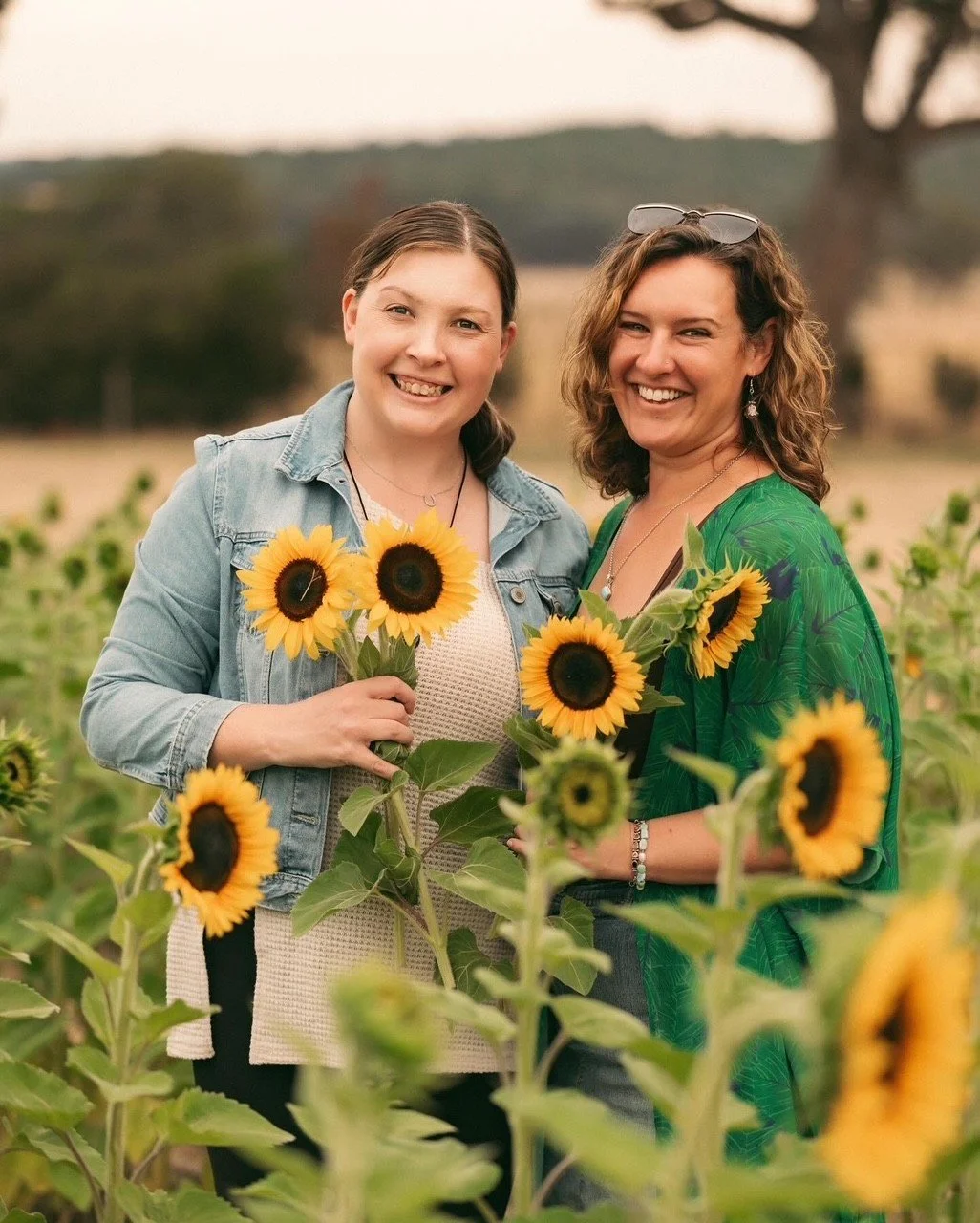 Two women smiling and holding sunflowers in a sunflower field during sunset.