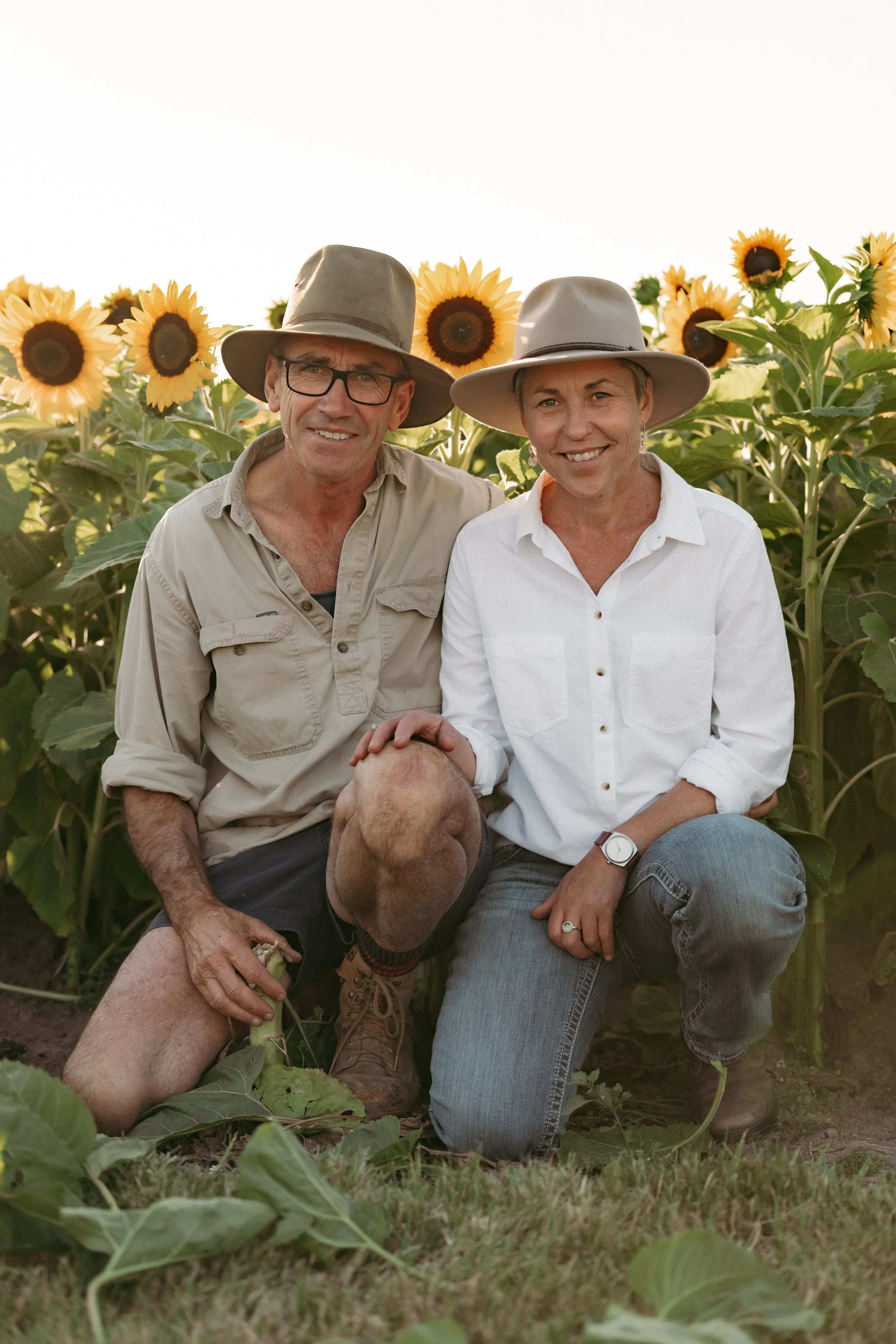 A man and a woman kneeling in a sunflower field, smiling at the camera, wearing hats and casual clothing.