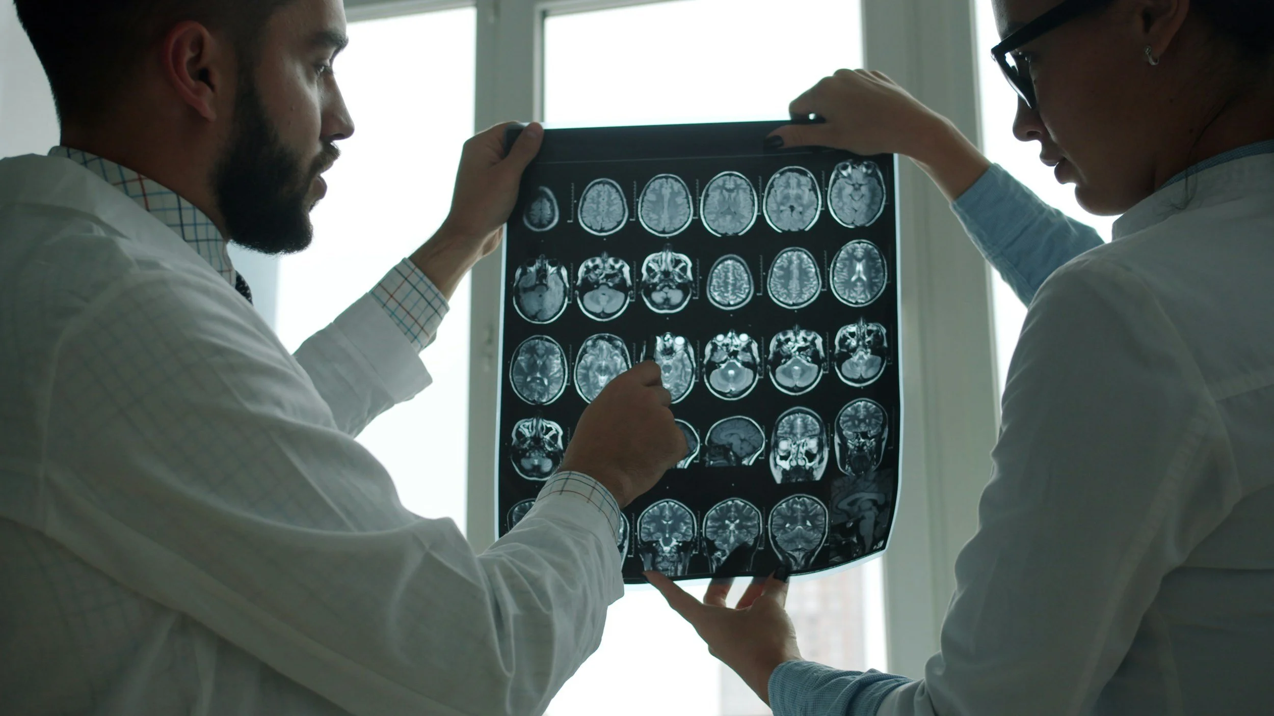 Two doctors examine an MRI brain scan together near a window.