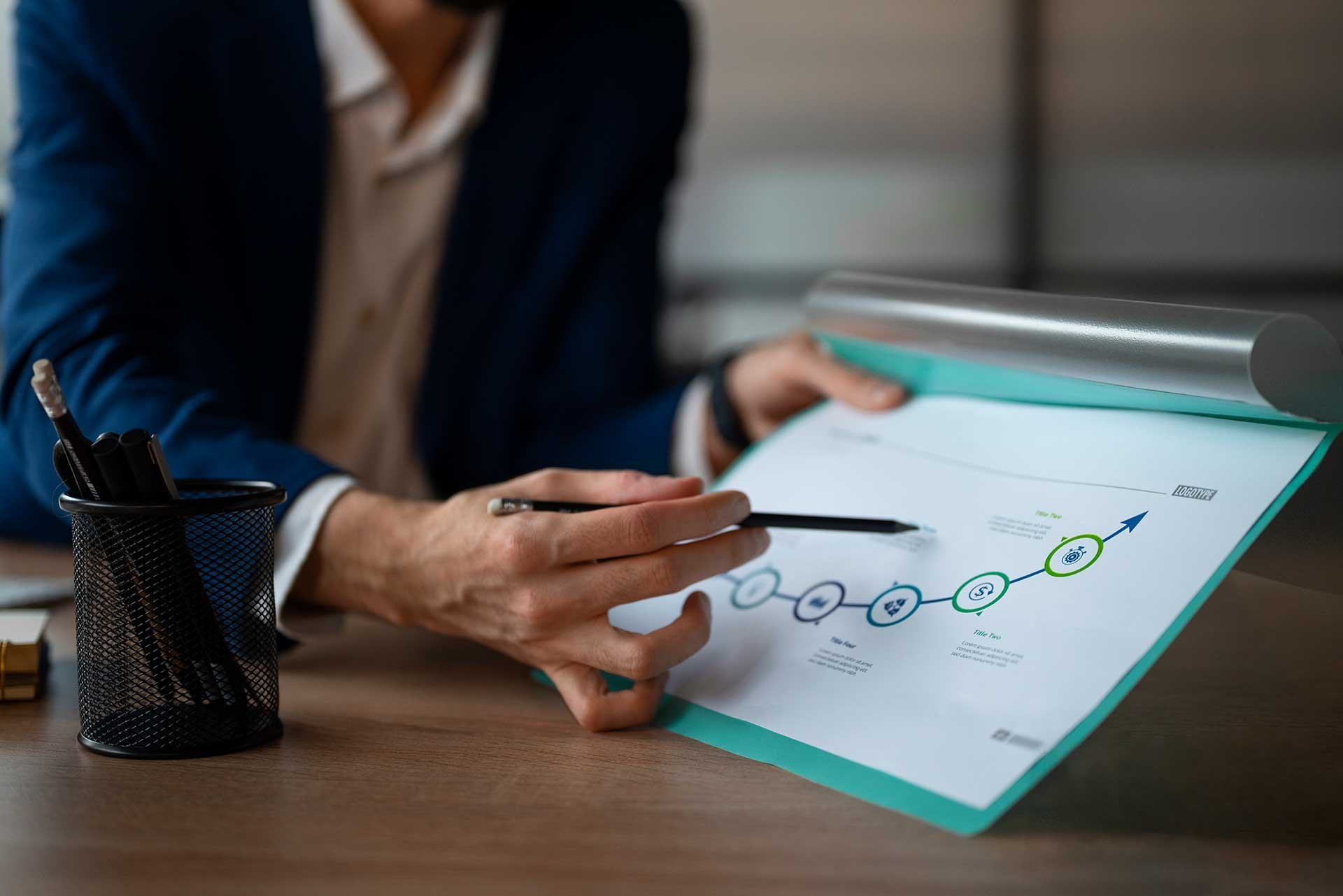 A person in business attire reviews a presentation chart on a clipboard, with a pen in hand, sitting at a desk with a pen holder.