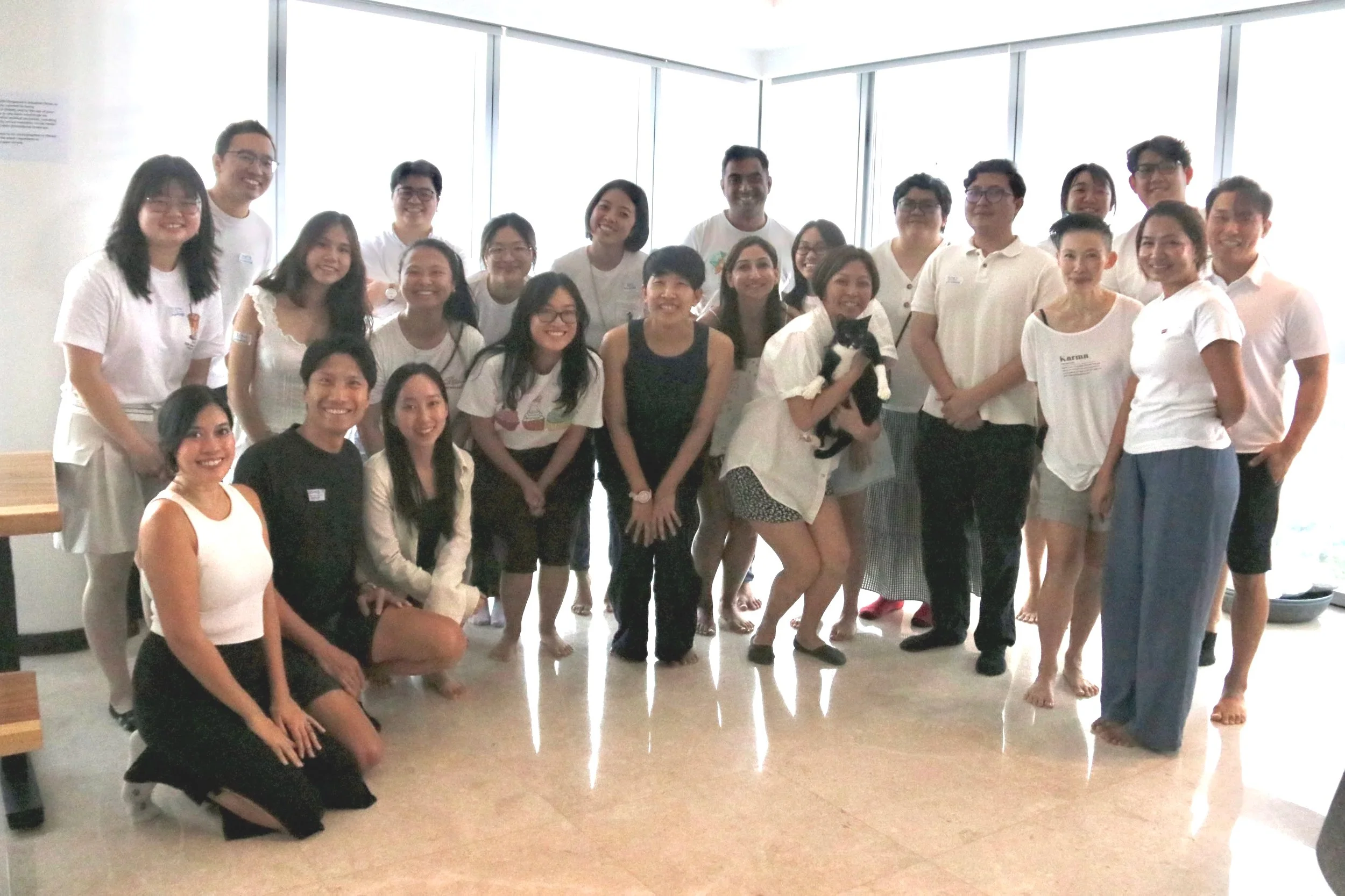 Group of people in casual attire, some kneeling and some standing, smiling for a photo in a bright room with large windows.
