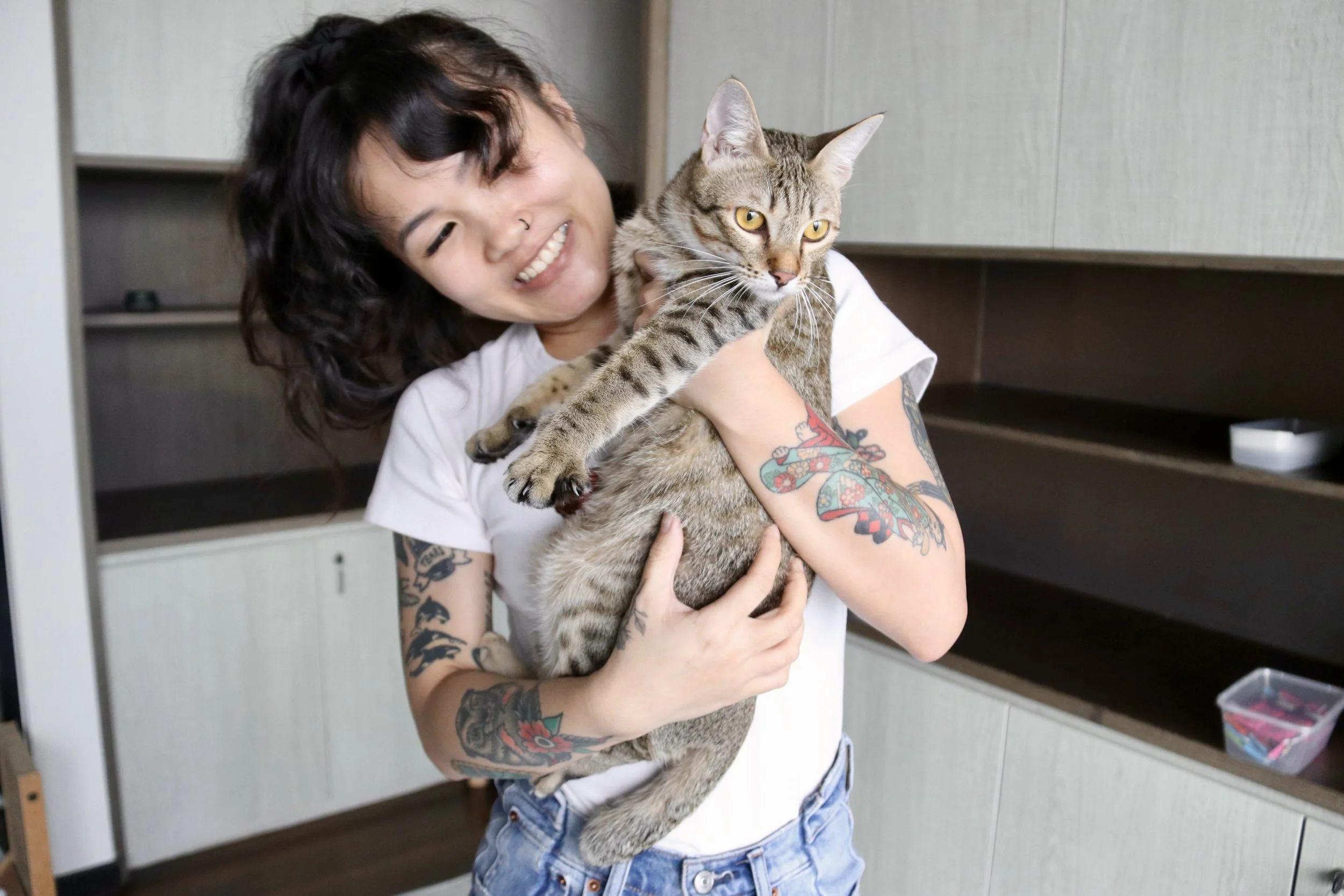 Smiling woman with tattoos holding a tabby cat indoors.