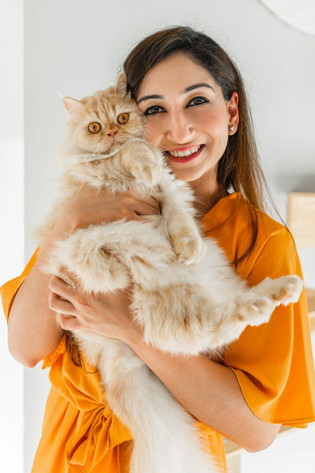 A woman wearing an orange dress holding a fluffy ginger cat with orange eyes, smiling at the camera.