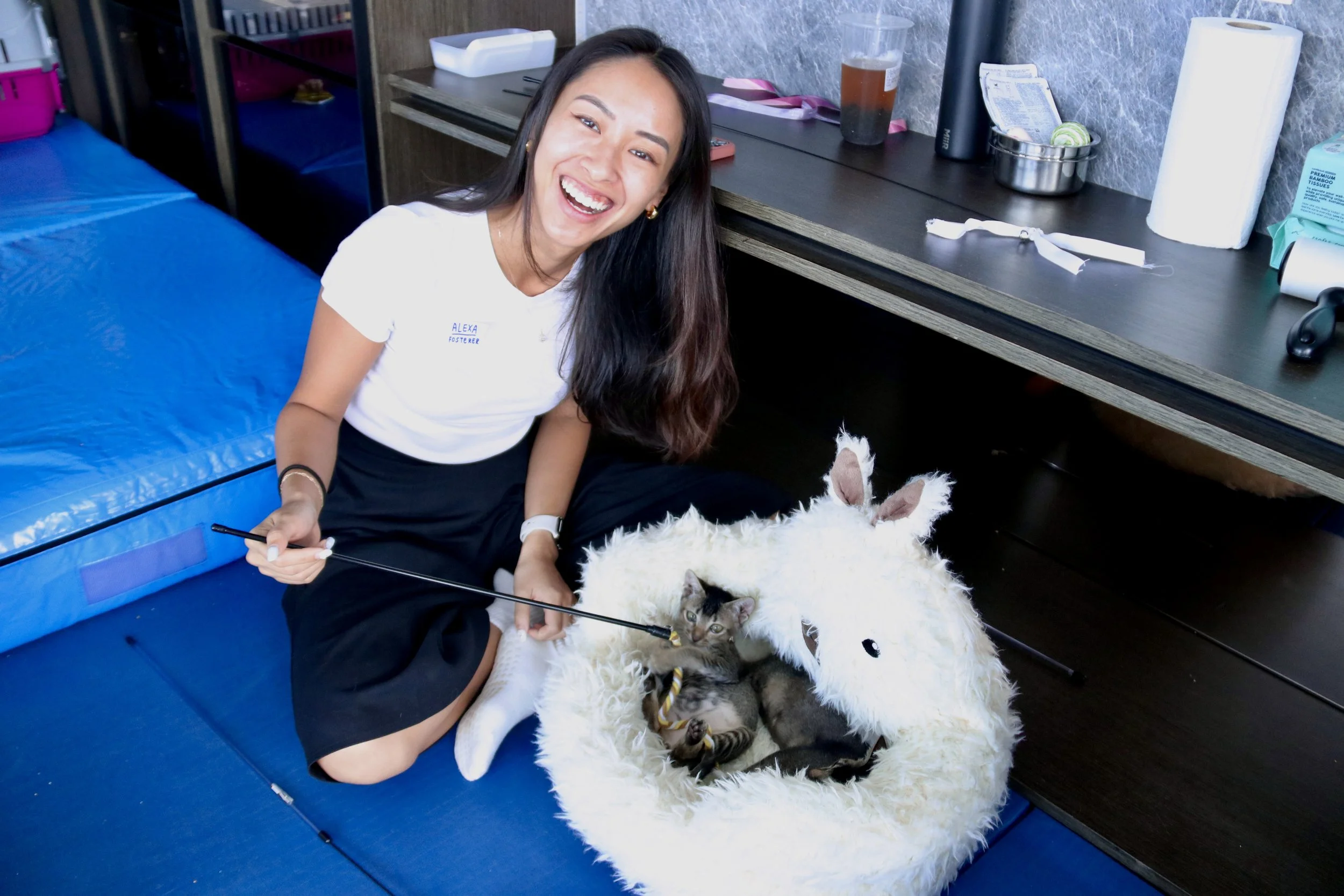 A woman with long dark hair, wearing a white t-shirt and black skirt, sitting on the floor and smiling while playing with a small kitten inside a plush white unicorn-shaped bed.