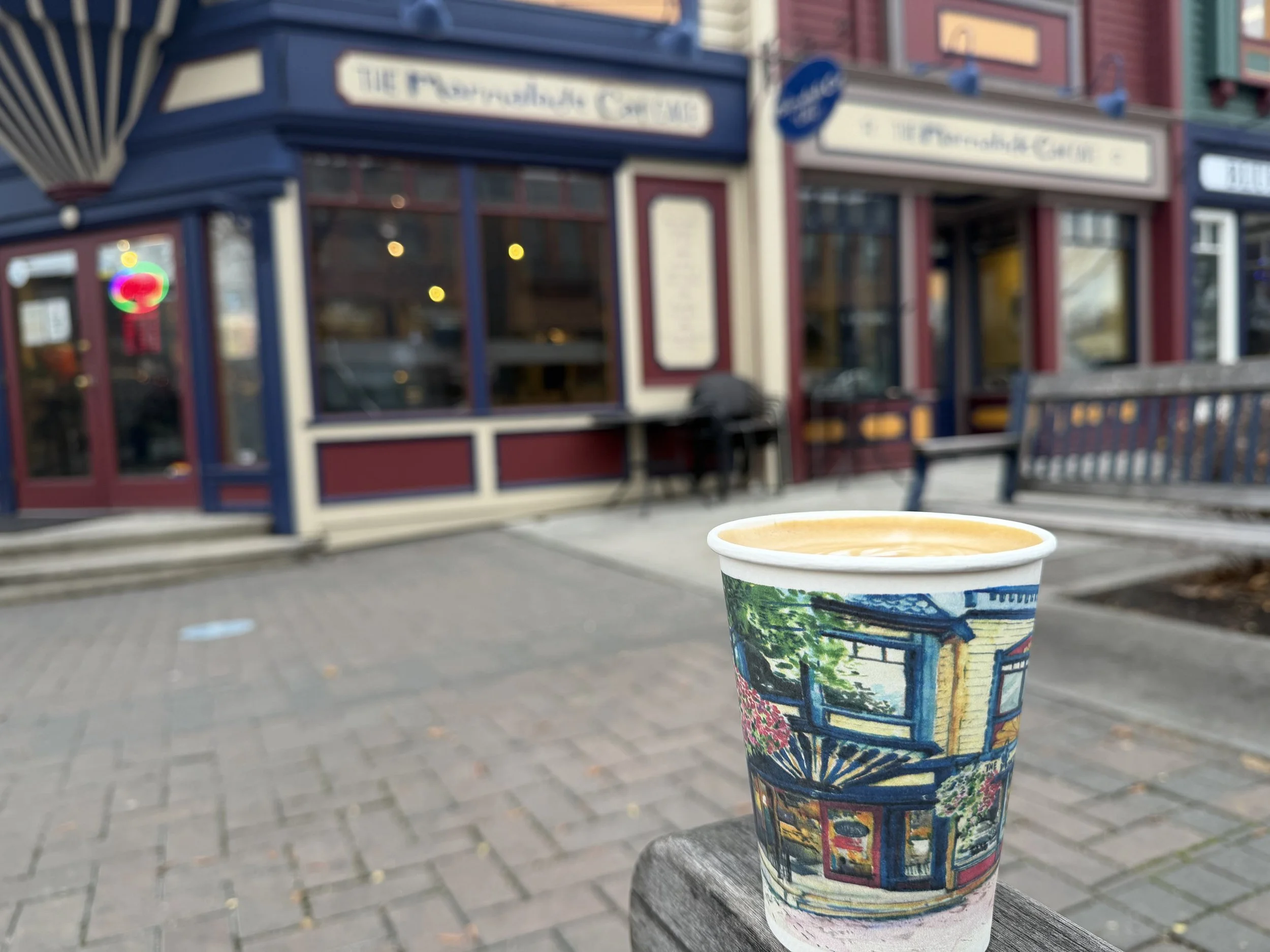 A cup of coffee rests on a table outside a restaurant or cafe, with a blurred storefront in the background.