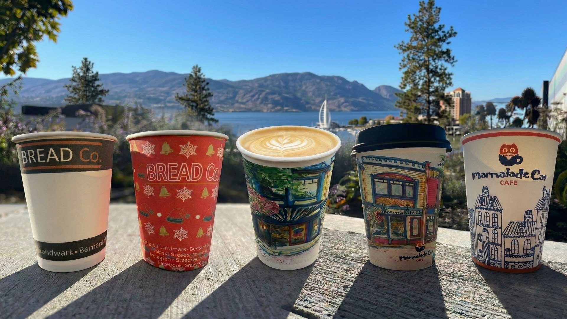 Five coffee cups on a wooden surface with mountain and lake view in the background. The cups have colorful designs and various branding.