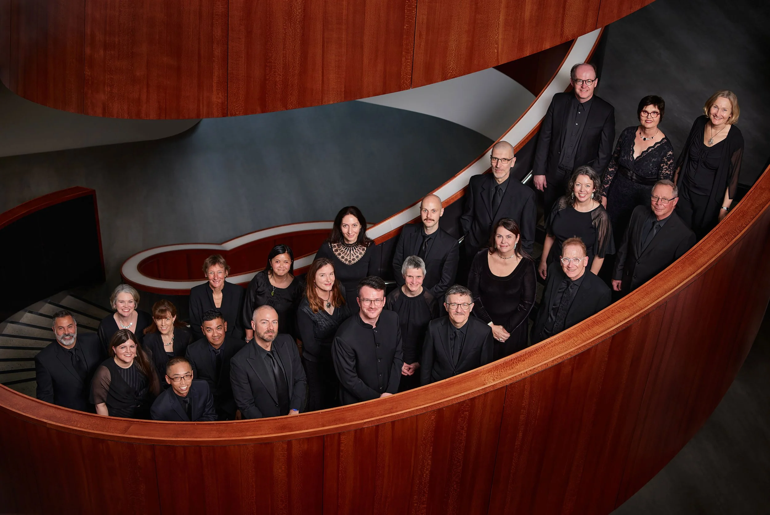 Sydney Chamber Choir and director Sam Alllchurch in formal black attire on a curved wooden staircase, with a dark background.