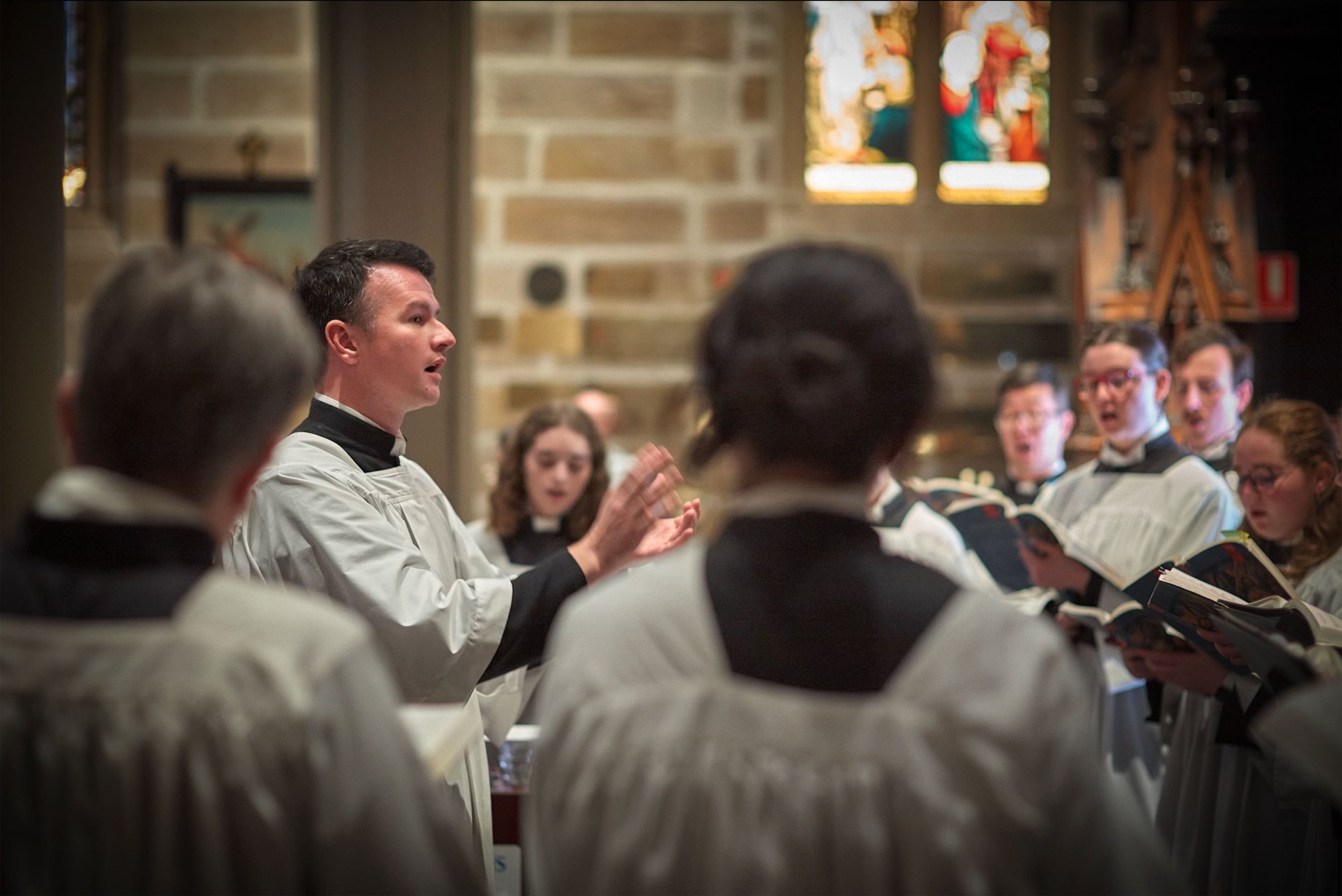 A choir conducted by Sam Allchurch, dressed in choir robes, with stained glass windows and brick walls in the background.