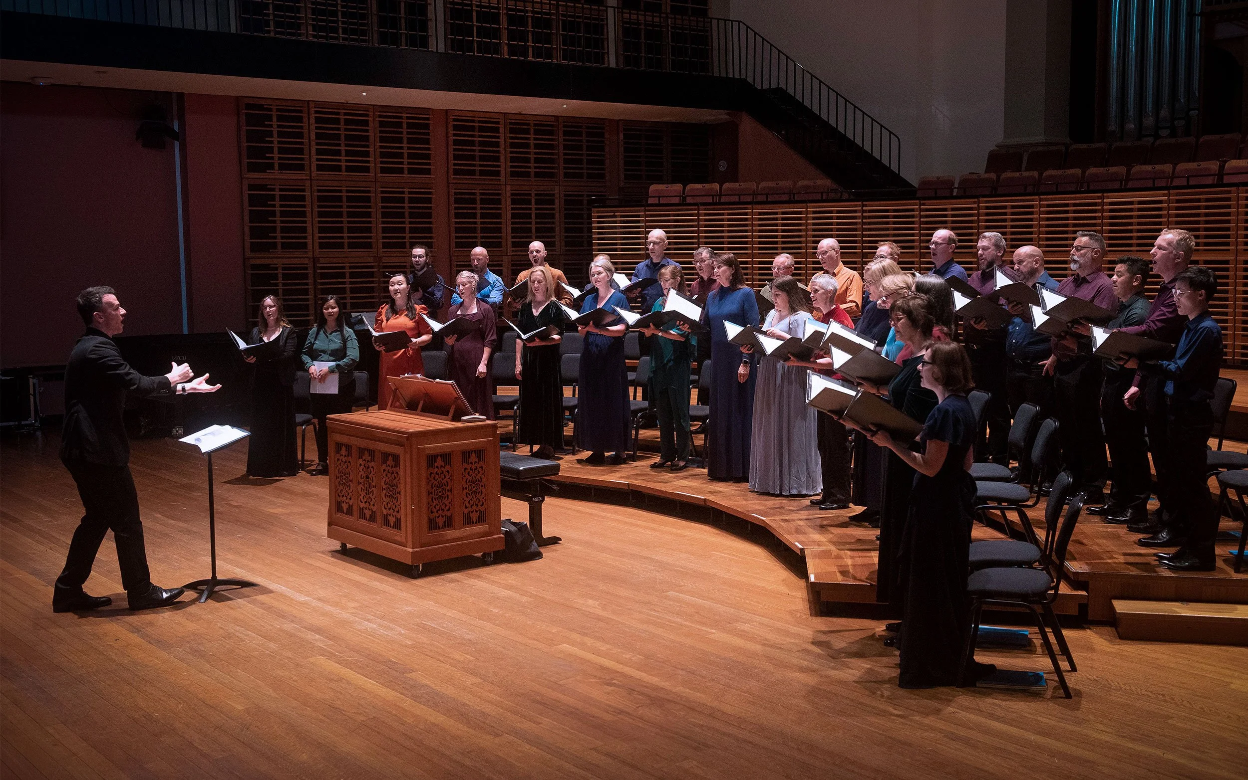 Sydney Chamber Choir, conducted by Sam Allchurch, performing on stage at Sydney Conservatorium of Music, a concert hall with wooden floors and seating.