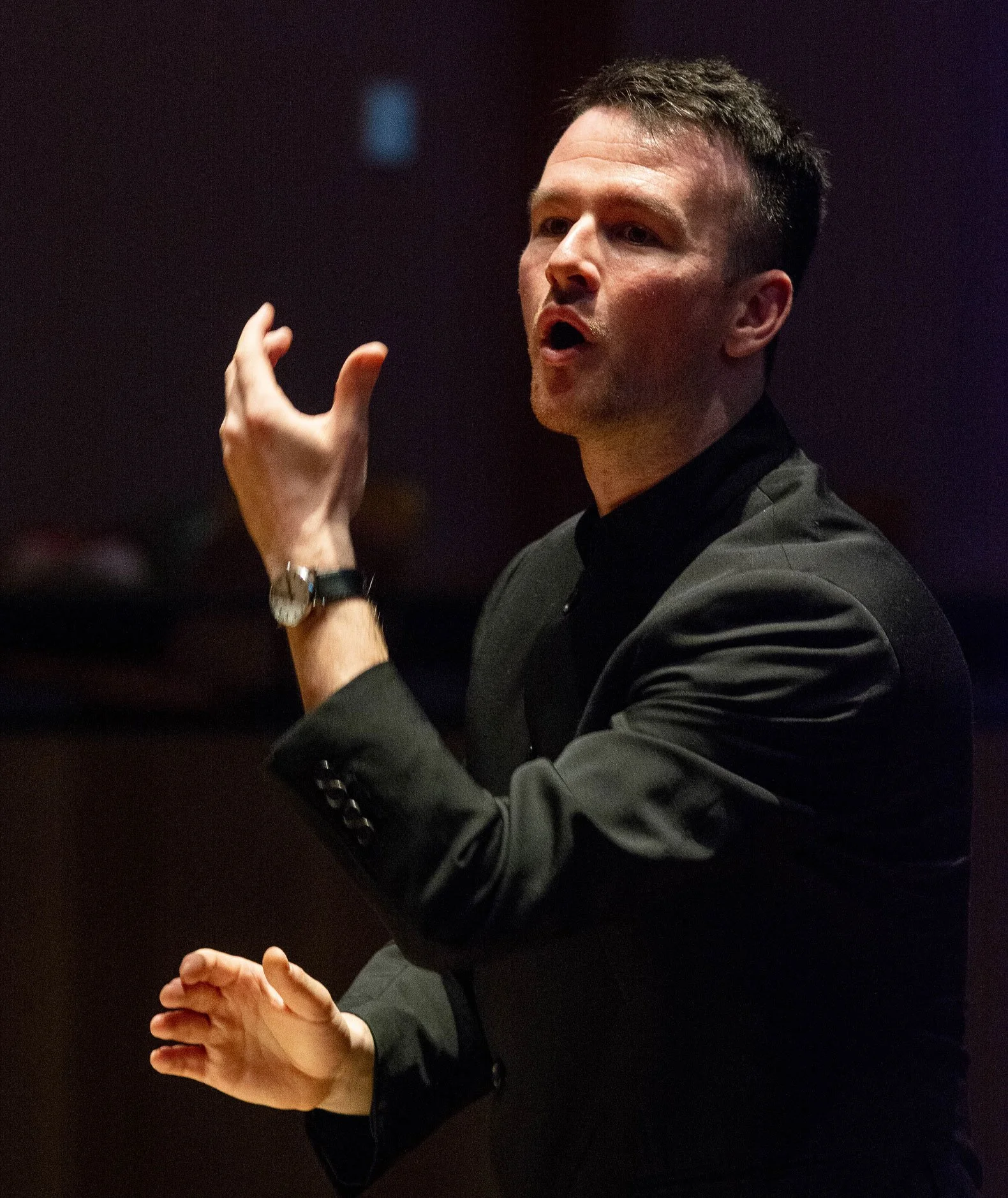 Sam Allchurch in a black shirt, conducting against a dark background.