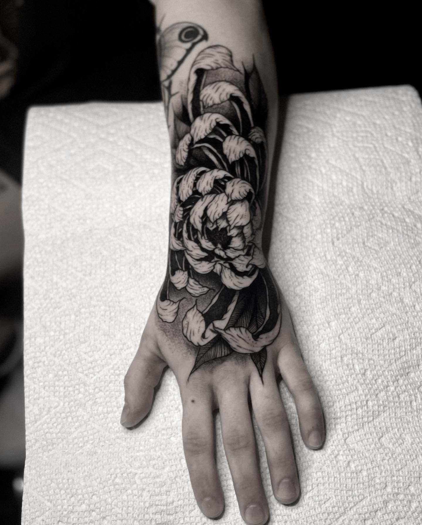 Black and gray tattoo of a large blooming rose with detailed petals and leaves on a person's forearm.
