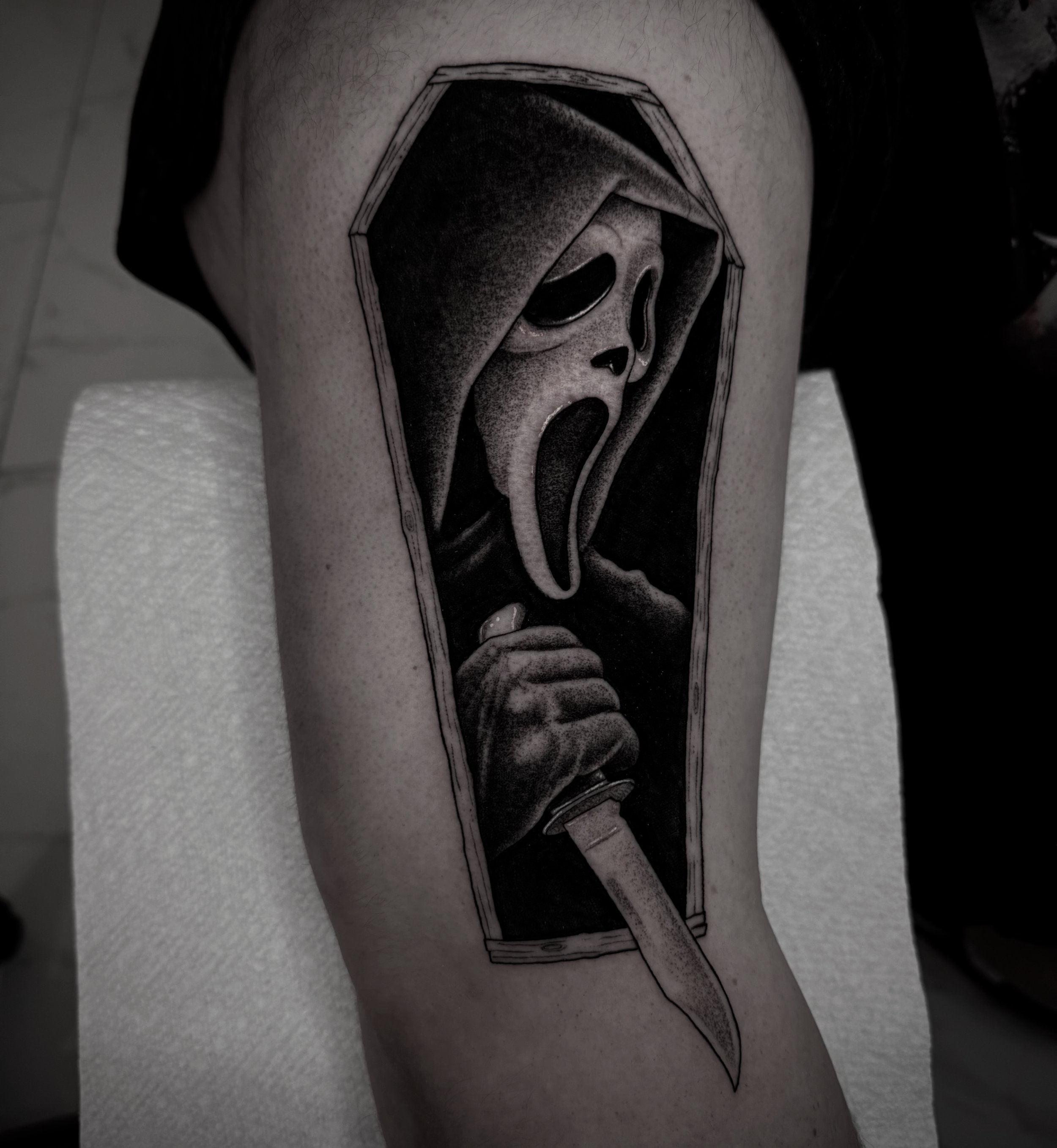 Tattoo of a person in a hood with a scream face, holding a bloody knife inside a coffin-shaped frame.