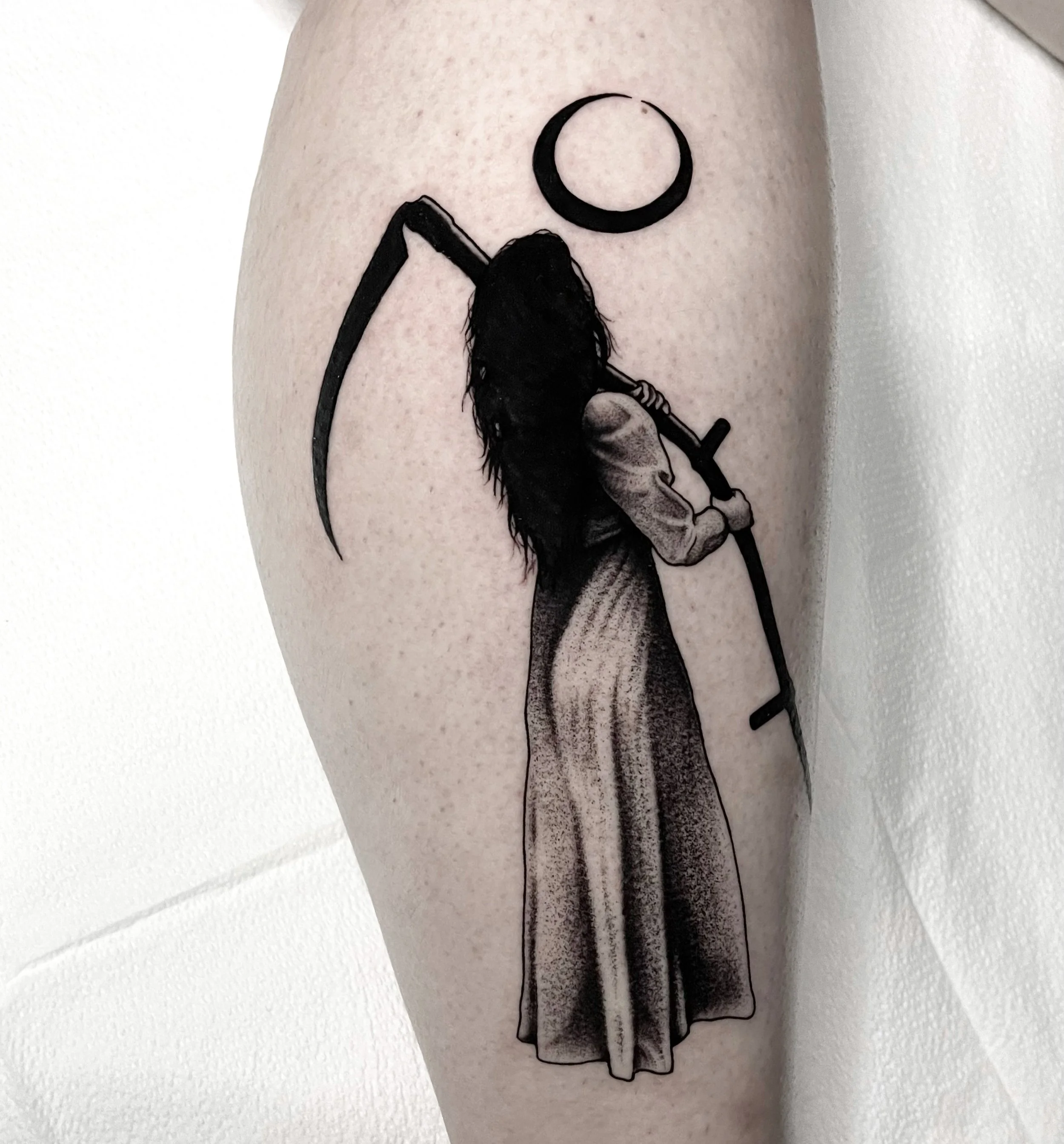 Tattoo of a woman with long hair holding a scythe, wearing a long skirt, with a crescent moon above her head.