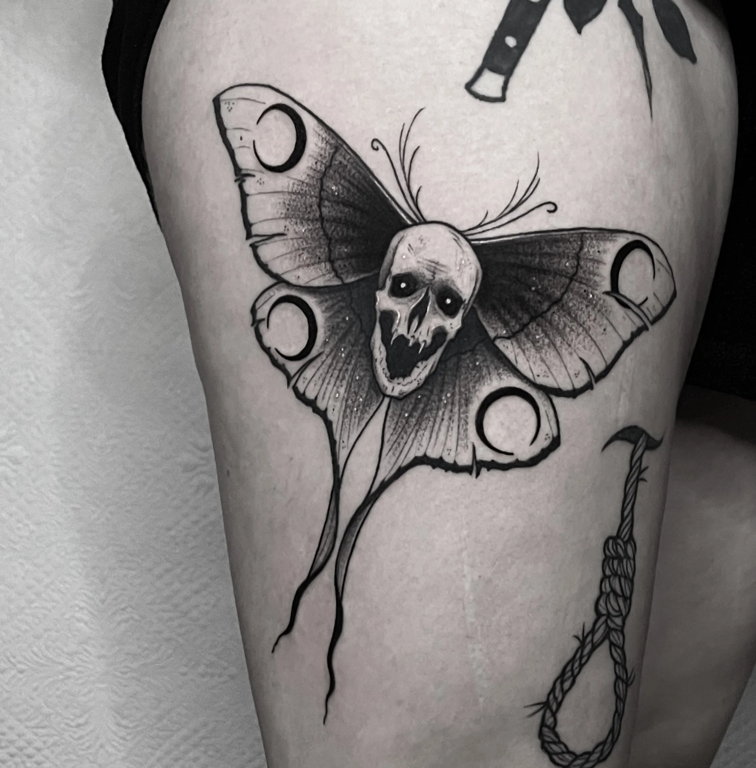 Black and white tattoo of a moth with a skull face on its body, surrounded by dark details and a hanging noose.