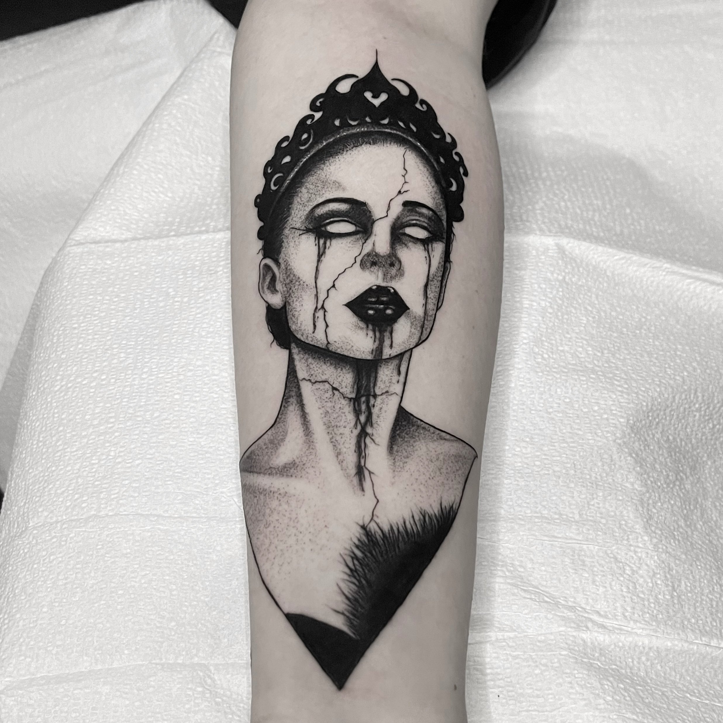 Black and gray tattoo of a woman with closed eyes, dark lipstick, tears running down her face, and a crown on her head, on a person's arm.