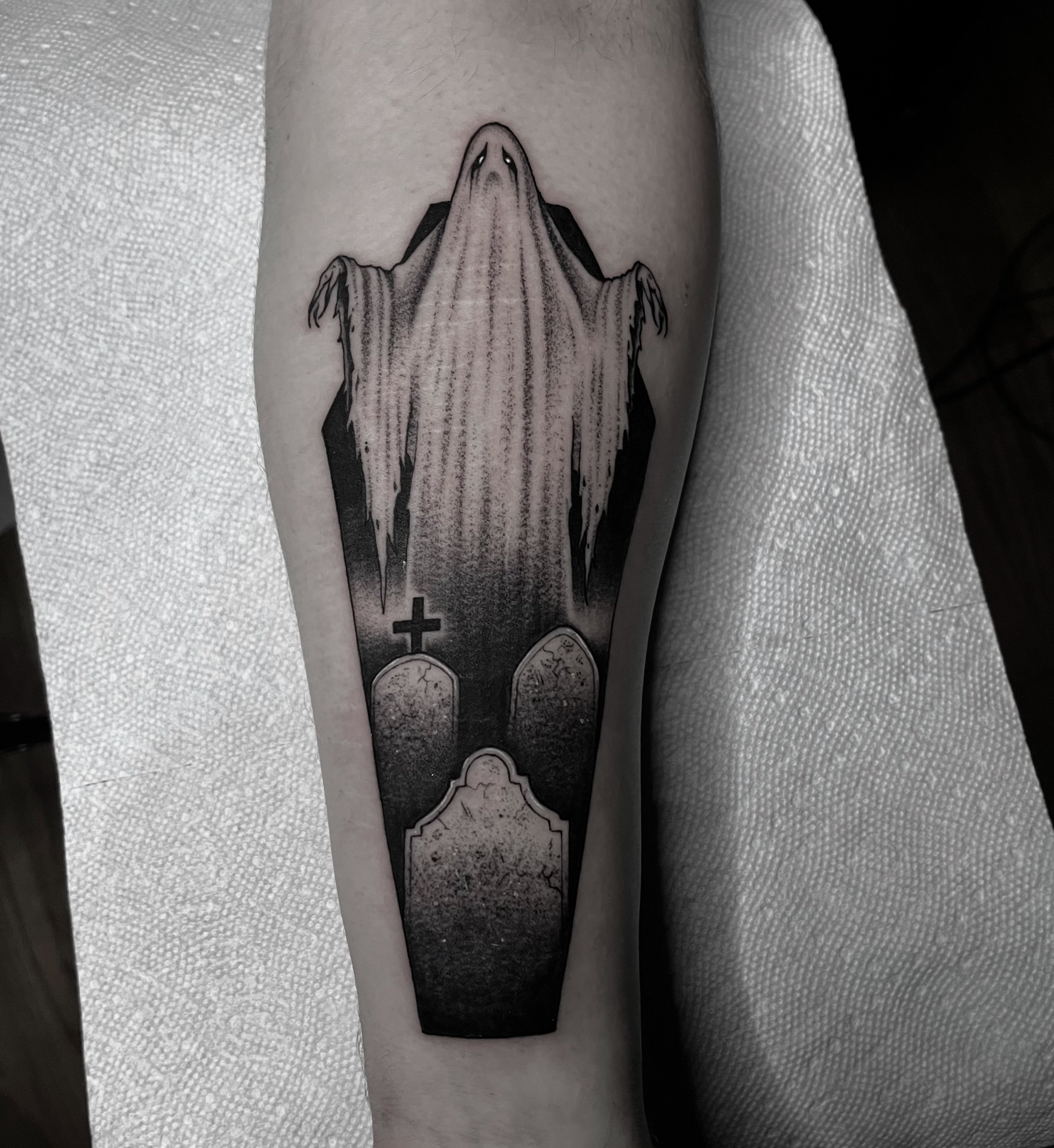 Black and gray tattoo of a ghostly figure with elongated face and draped cloak, standing above three gravestones, one with a cross, on a person's arm.