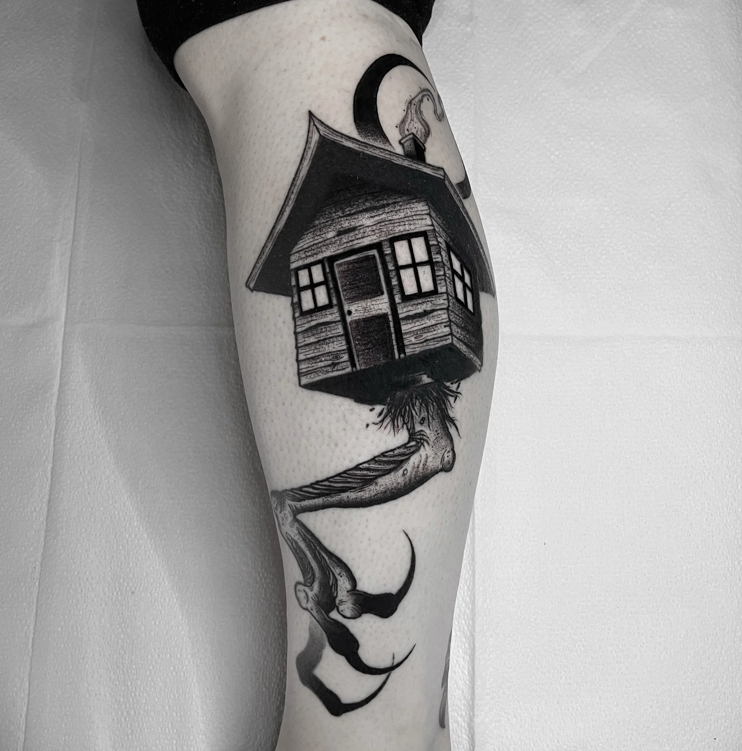 Black and gray tattoo of a small wooden house on stilts with a staircase underneath, and a crescent moon above, on a person's arm.