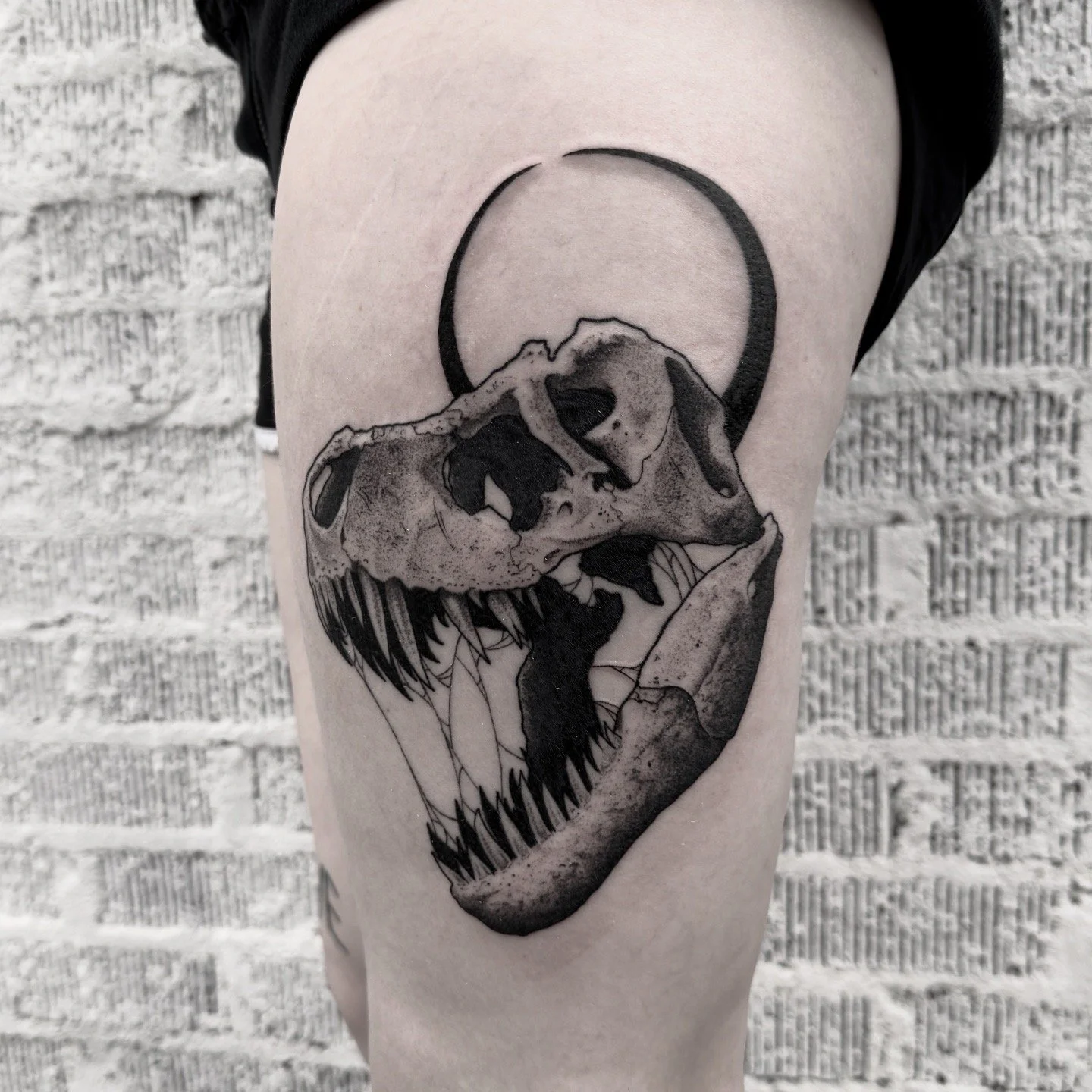 Tattoo of a T-Rex skull with an eclipse circle above it, located on a person's upper arm.