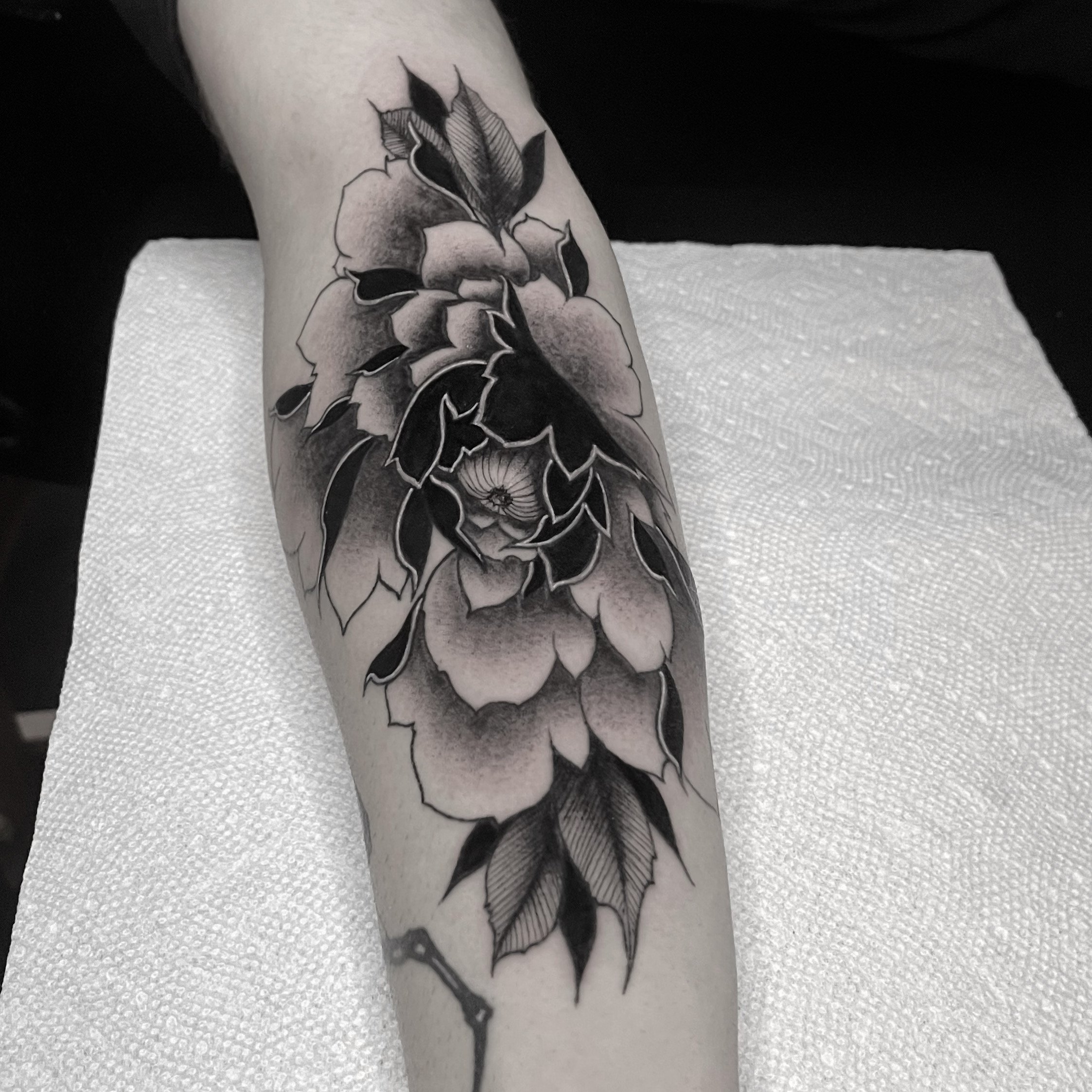 Black and gray floral tattoo on forearm featuring a large blooming flower with detailed petals and leaves.