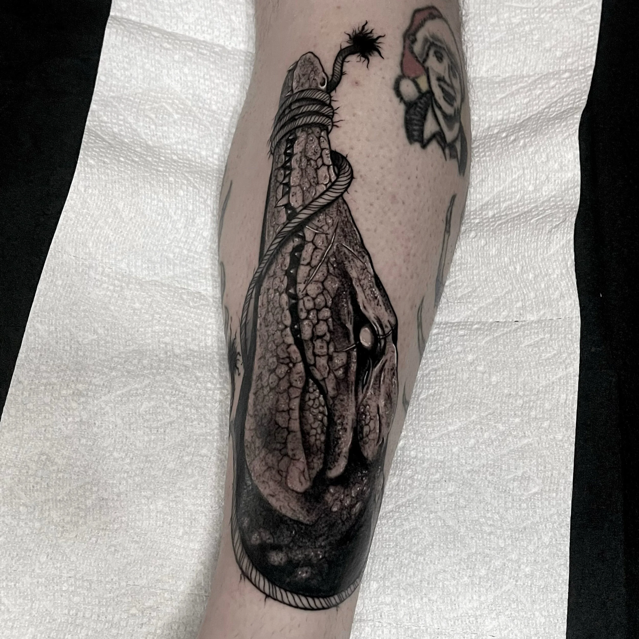 Black and gray tattoo of a dinosaur head on a person's forearm, with a smaller tattoo of a colorful clown face nearby.