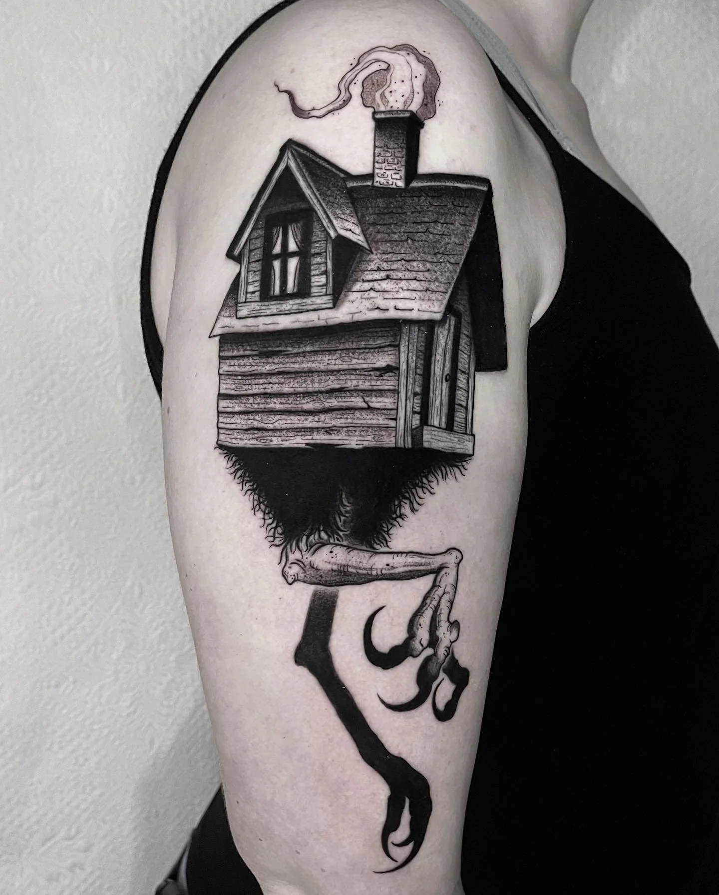 Tattoo of a haunted house with a chimney emitting smoke, perched on a bird's wing, with a claw extending downward.