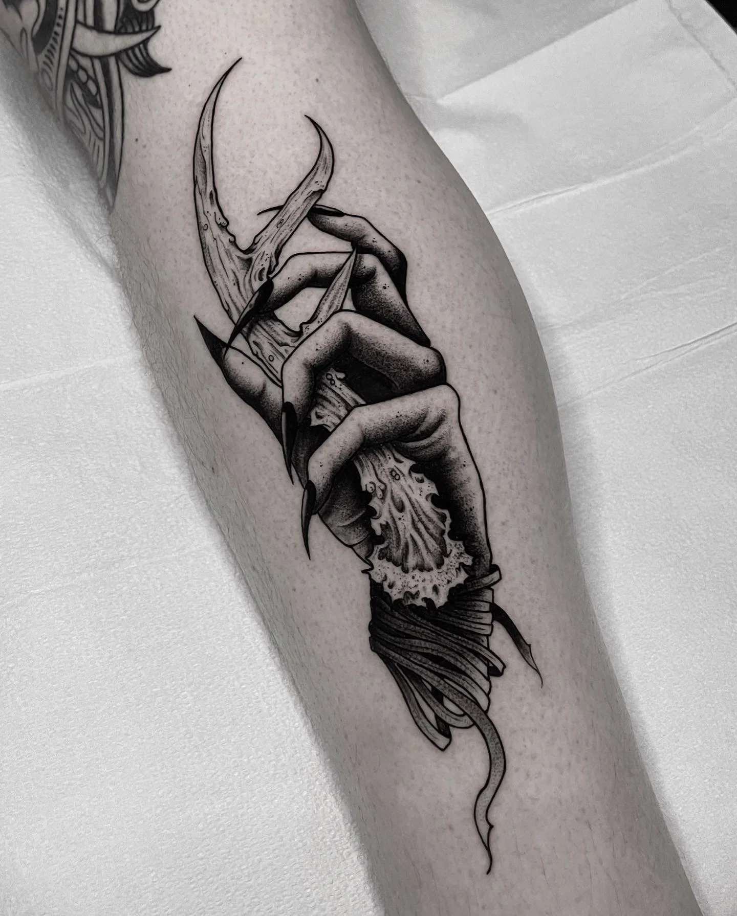 Black and gray tattoo of a hand with long, pointed nails gripping a jagged, broken object, wrapped with a ribbon or band at the wrist.