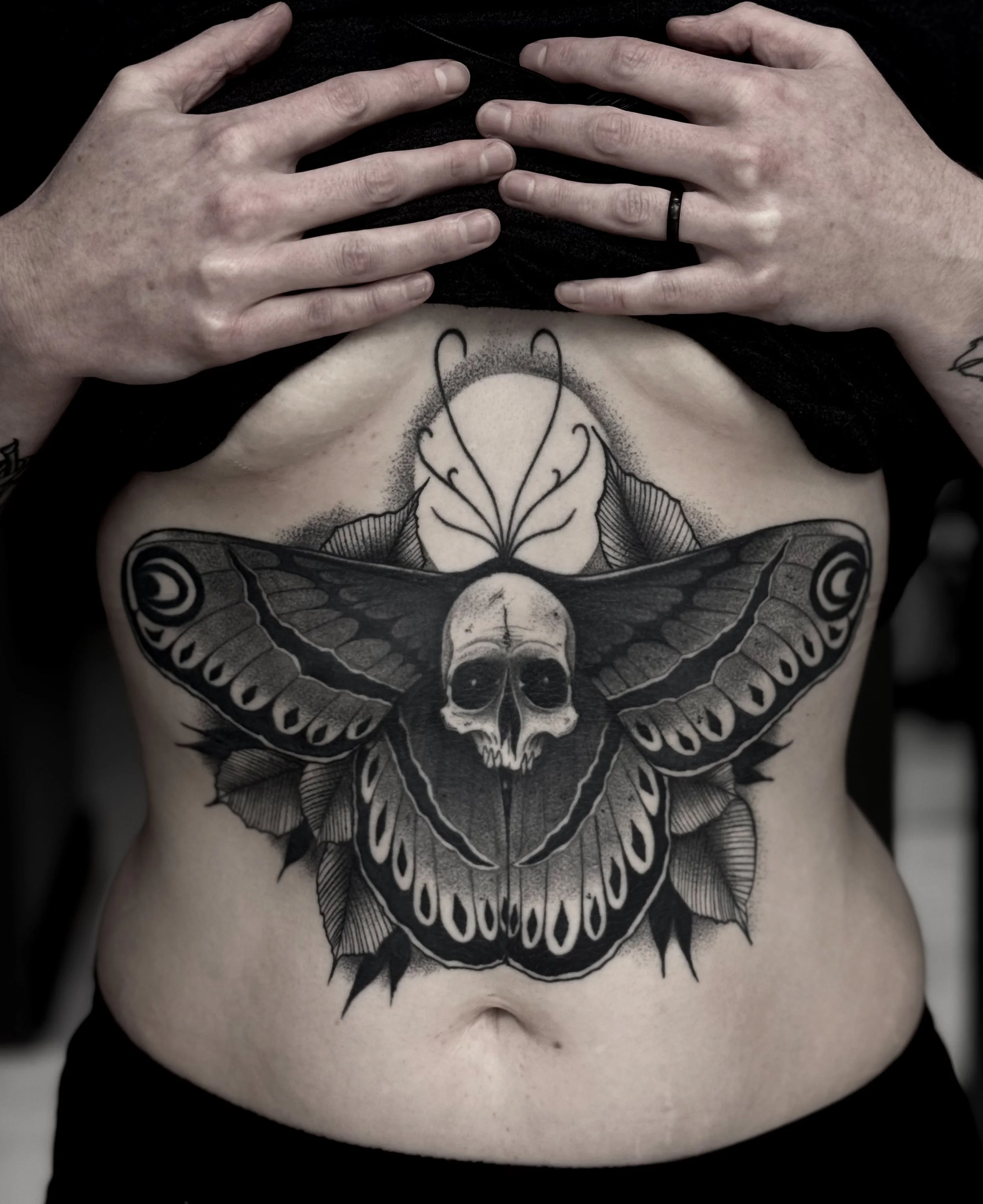 Black and gray tattoo of a moth with a skull on its body, surrounded by leaves, on a person's stomach. Two hands are resting on the person's chest.