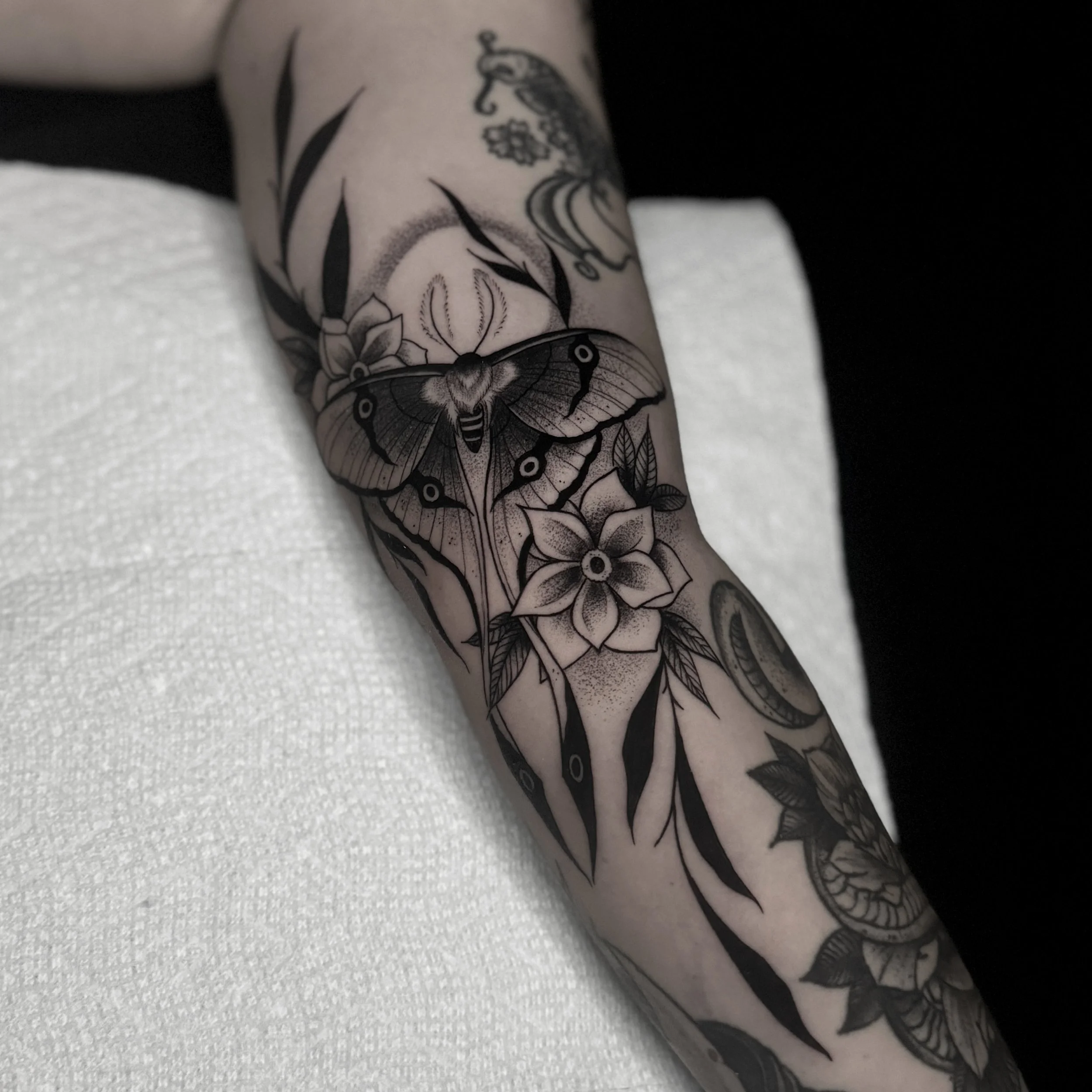 Black and gray tattoo of a moth, flowers, and leaves on a person's arm.