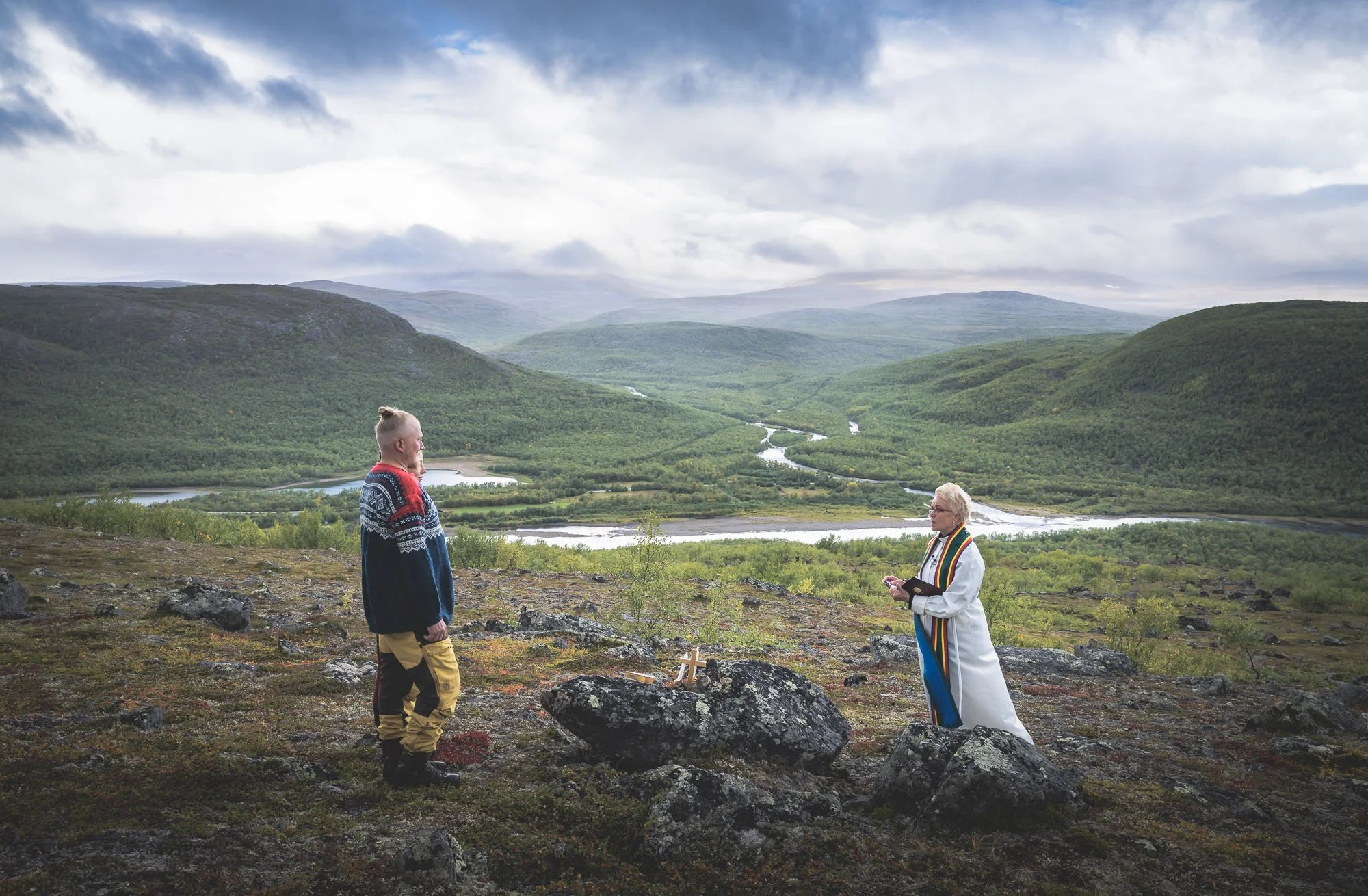 A man and a woman standing on a rocky hillside during a ceremony with a mountainous landscape with rivers and forests in the background.