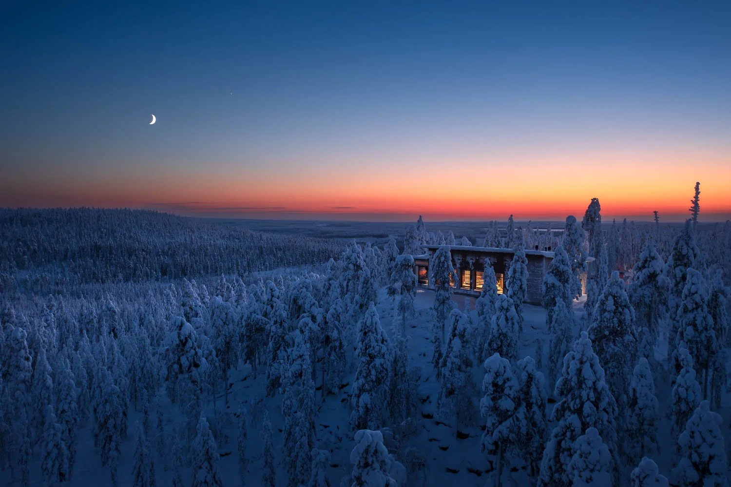 Snow-covered forest with a modern cabin emitting warm light against a colorful sunset and a crescent moon in the sky.