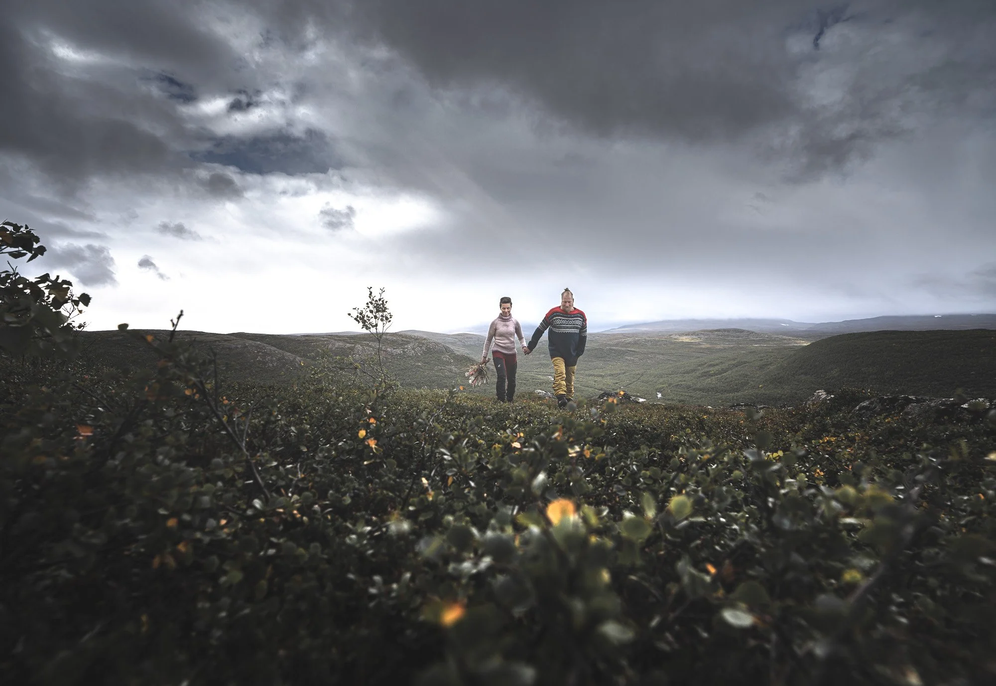 A couple hiking hand in hand through a rugged, hilly landscape under a cloudy sky, with vegetation in the foreground.