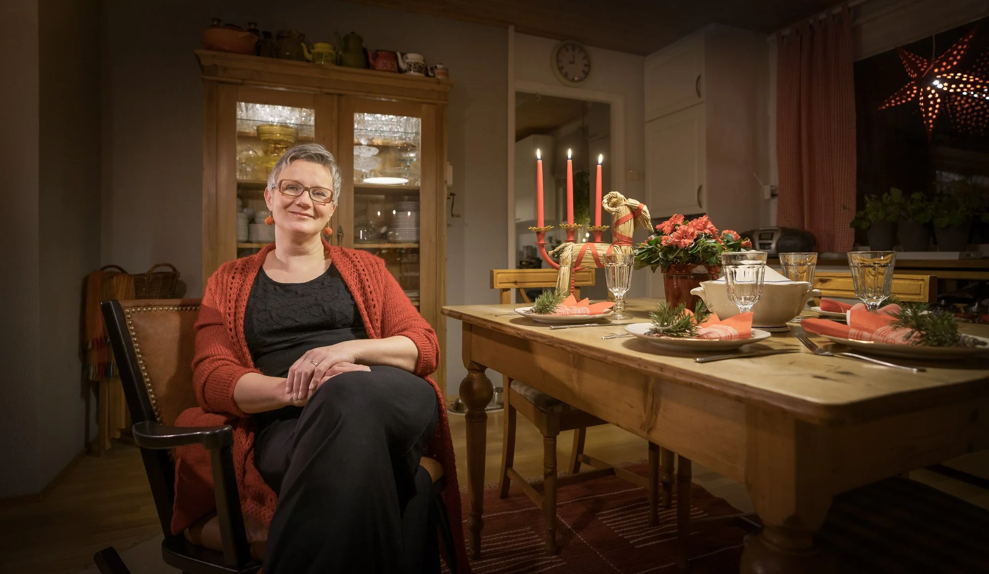 A woman with glasses and short gray hair, wearing a black dress and a red knitted cardigan, sitting in a dining room with a festive table set with flowers, candles, and holiday decorations.