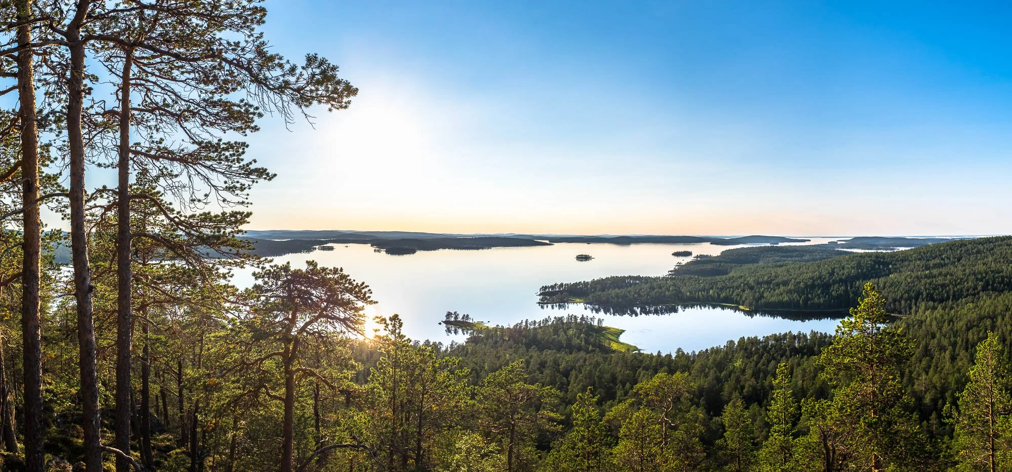 A scenic view of a lake Inarijärvi surrounded by dense green forest under a clear blue sky, with the sun near the horizon reflecting on the water.