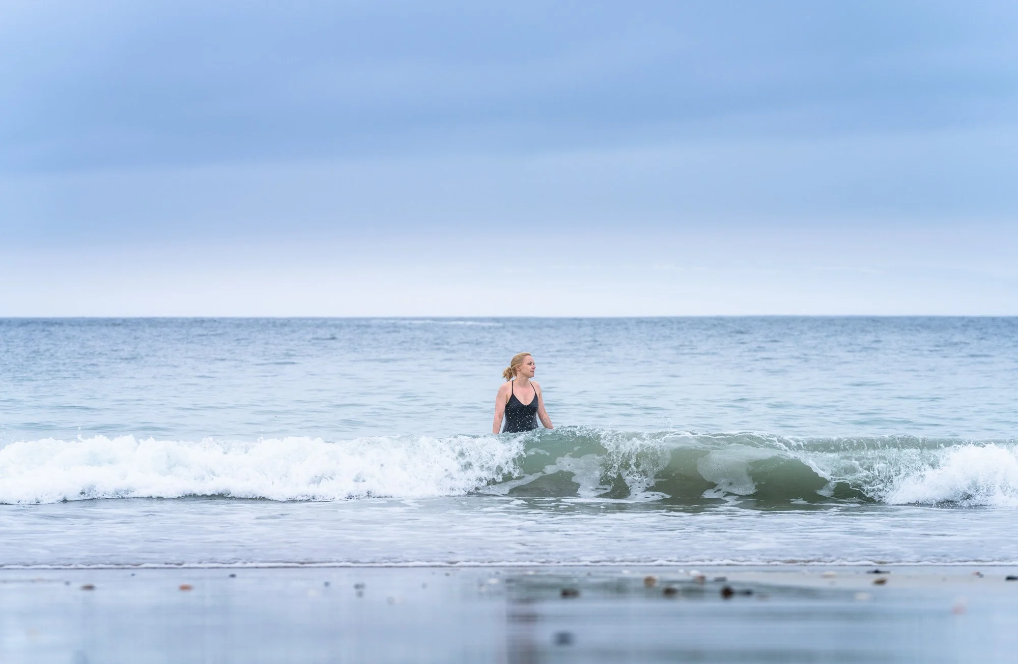 A woman in a swimsuit standing in the ocean waves at the beach under a cloudy sky.