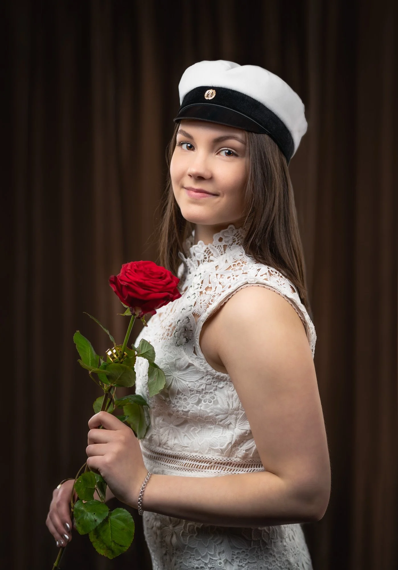 A young woman with long brown hair wearing a white lace sleeveless dress and a white and black hat, holding a red rose, standing in front of a dark brown curtain.