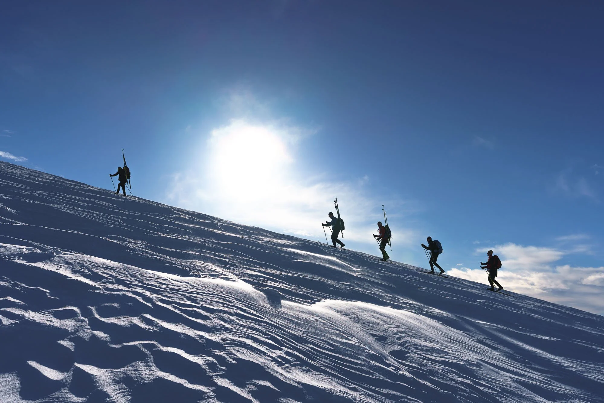 Five skiers ascending a snowy mountain slope under a blue sky with the sun shining behind them.