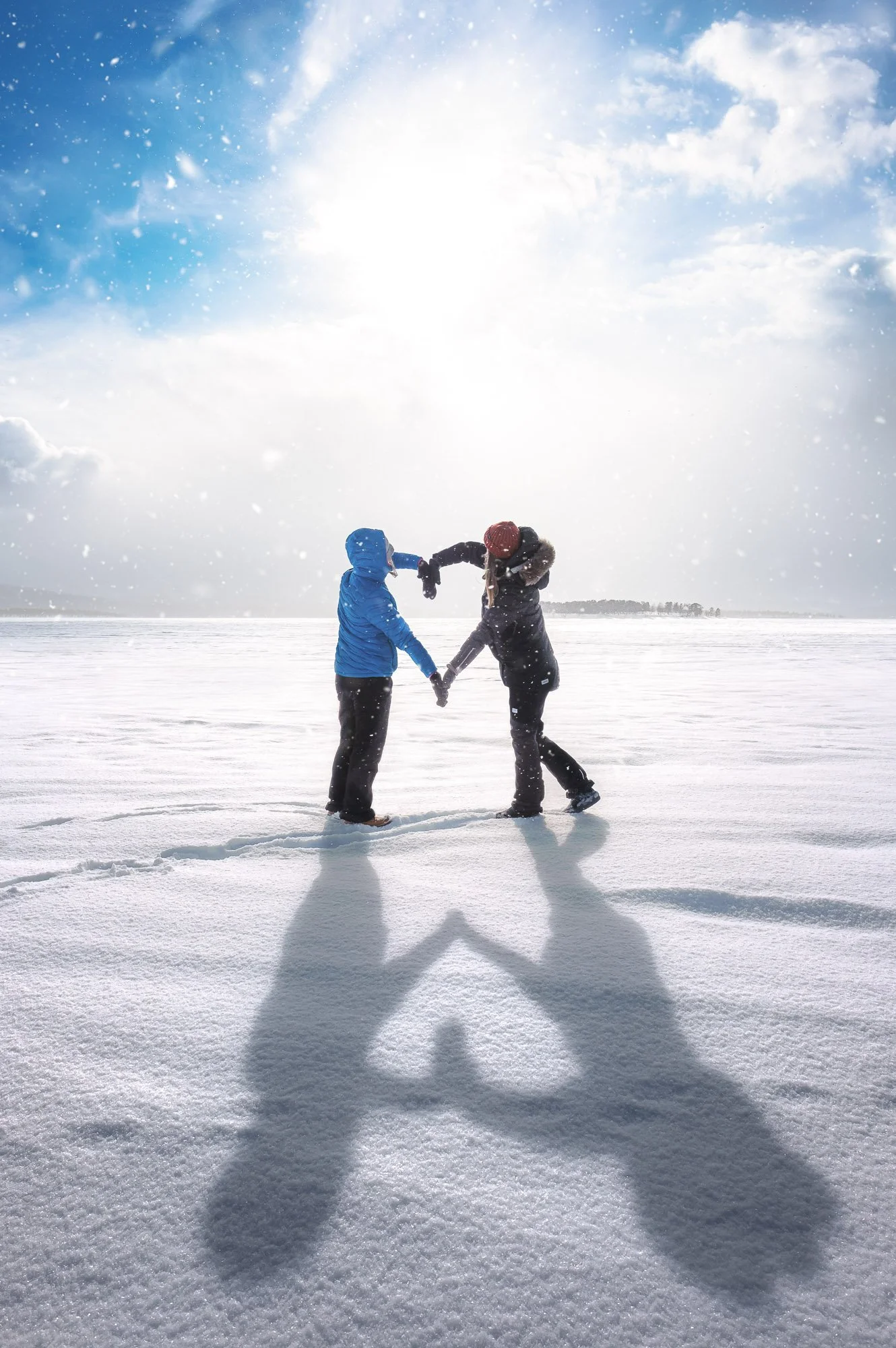 Two people holding hands and making a heart shape with their shadows on the snow, standing on a snowy landscape with a bright sun in the sky and snow falling.