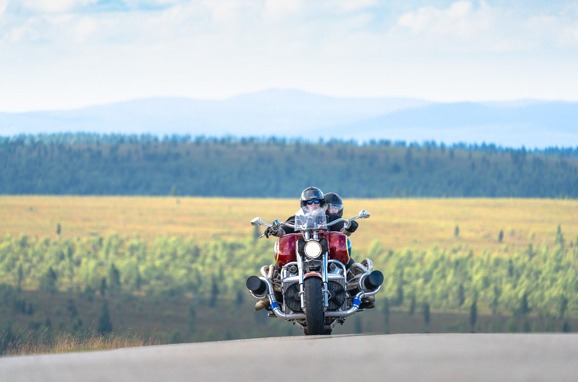 A person riding a red motorcycle on a rural road with green fields and hills in the background.