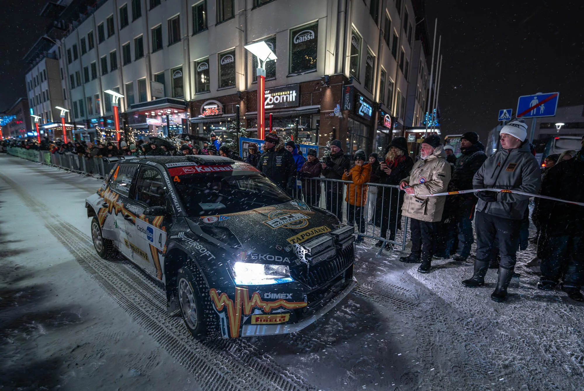A rally car on a snowy street with spectators behind a barrier, surrounded by buildings with illuminated signs at night.