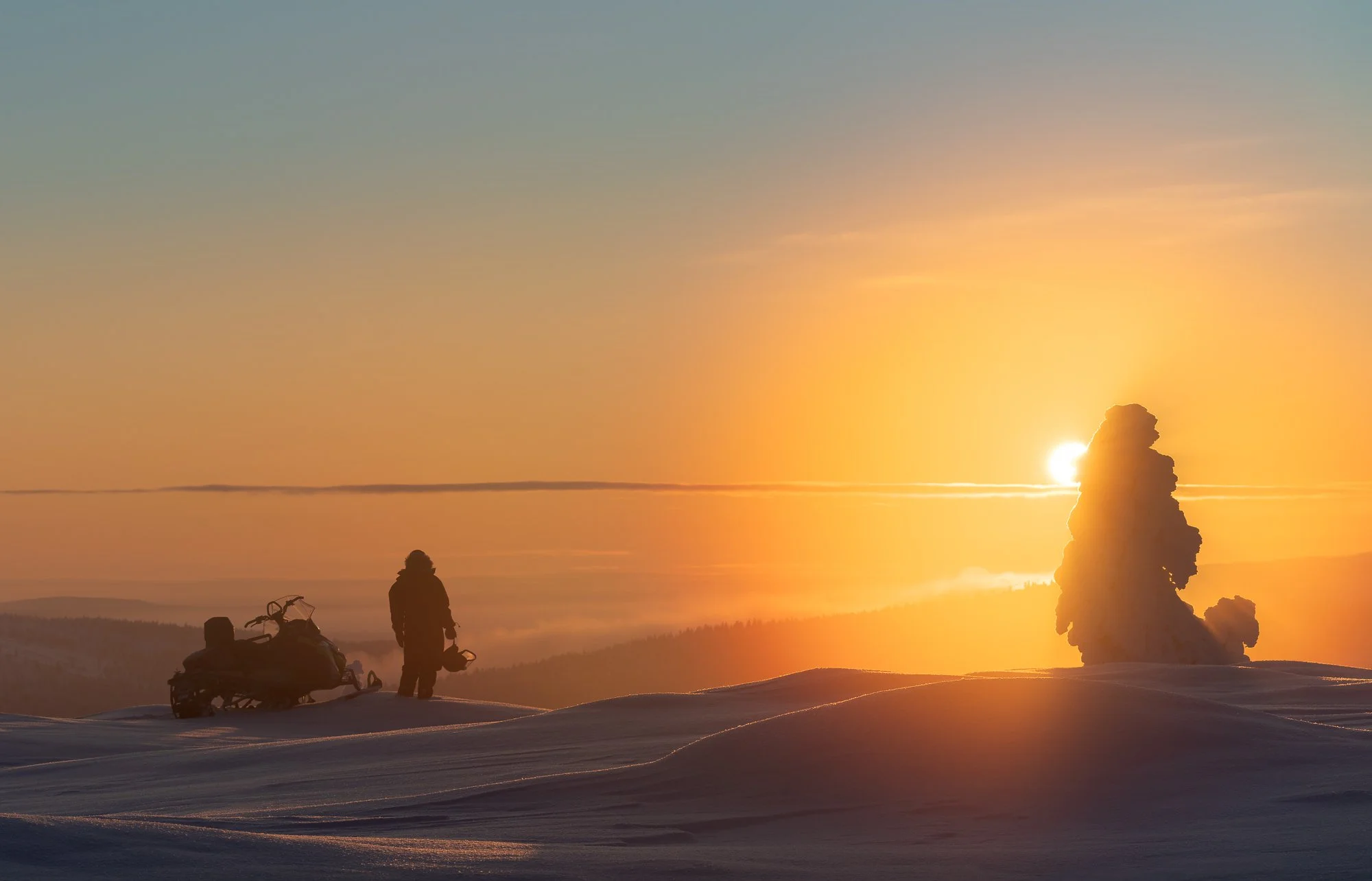 A person with a snowmobile standing on snowy terrain during sunset, with a large ice formation in the background.
