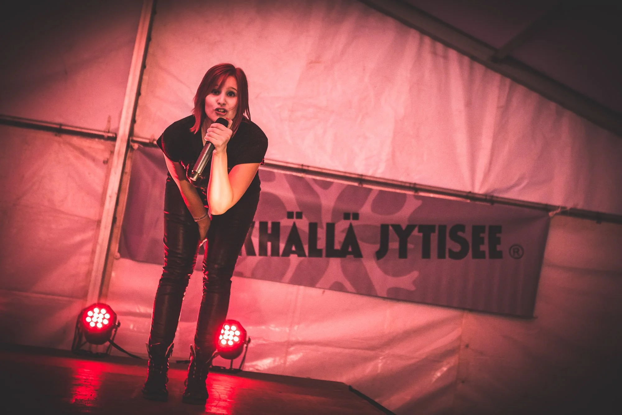 A woman with red hair singing into a microphone on stage, with red stage lights and a banner with the word 'KÄLLÄ JYTISEE' behind her.