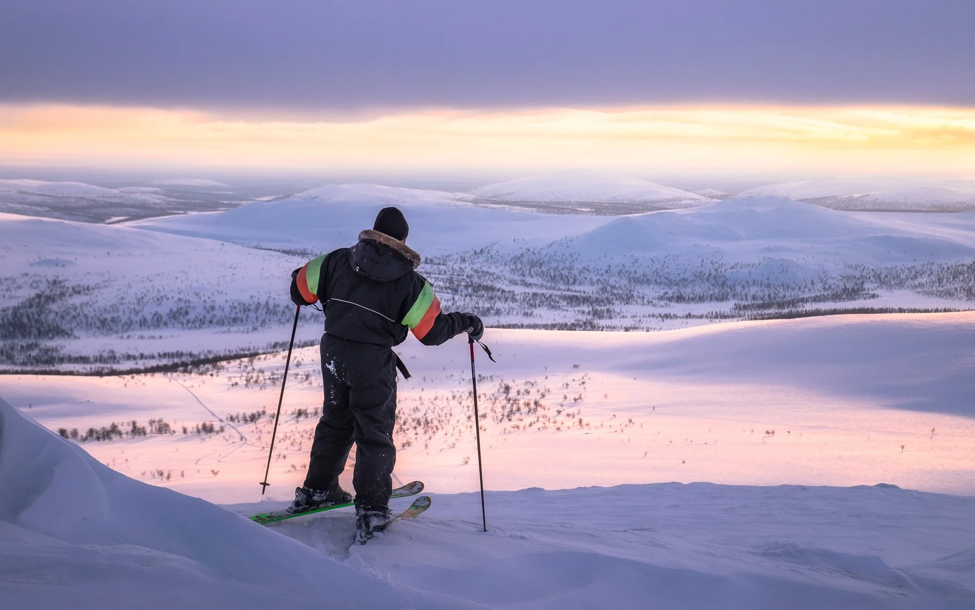 A person dressed in winter clothing skiing on snow-covered terrain, overlooking a vast, snowy landscape with hills and trees under a colorful sky at sunset or sunrise.